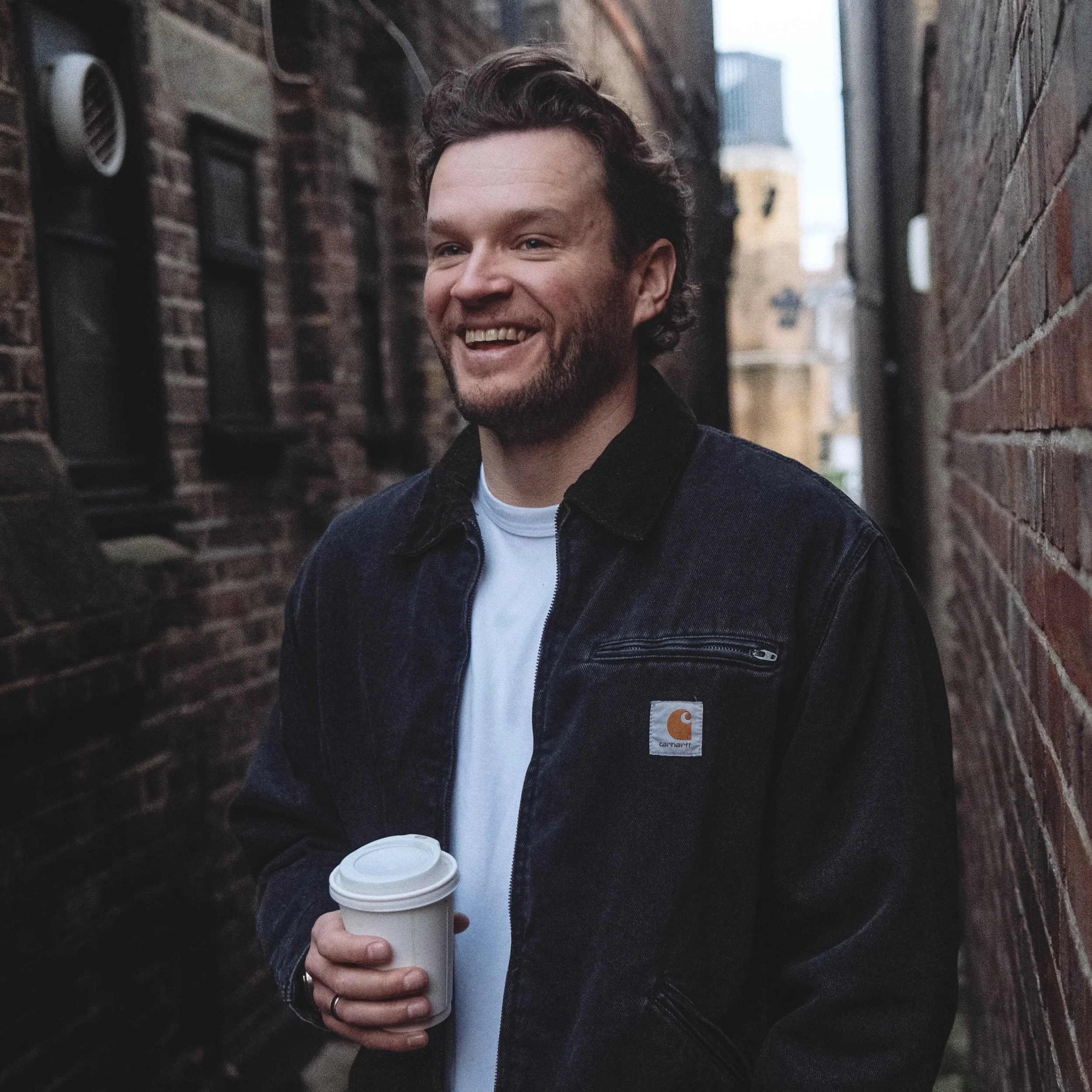 A man with a beard and wavy hair smiling, wearing a black jacket, holding a disposable coffee cup, standing between brick walls outdoors.