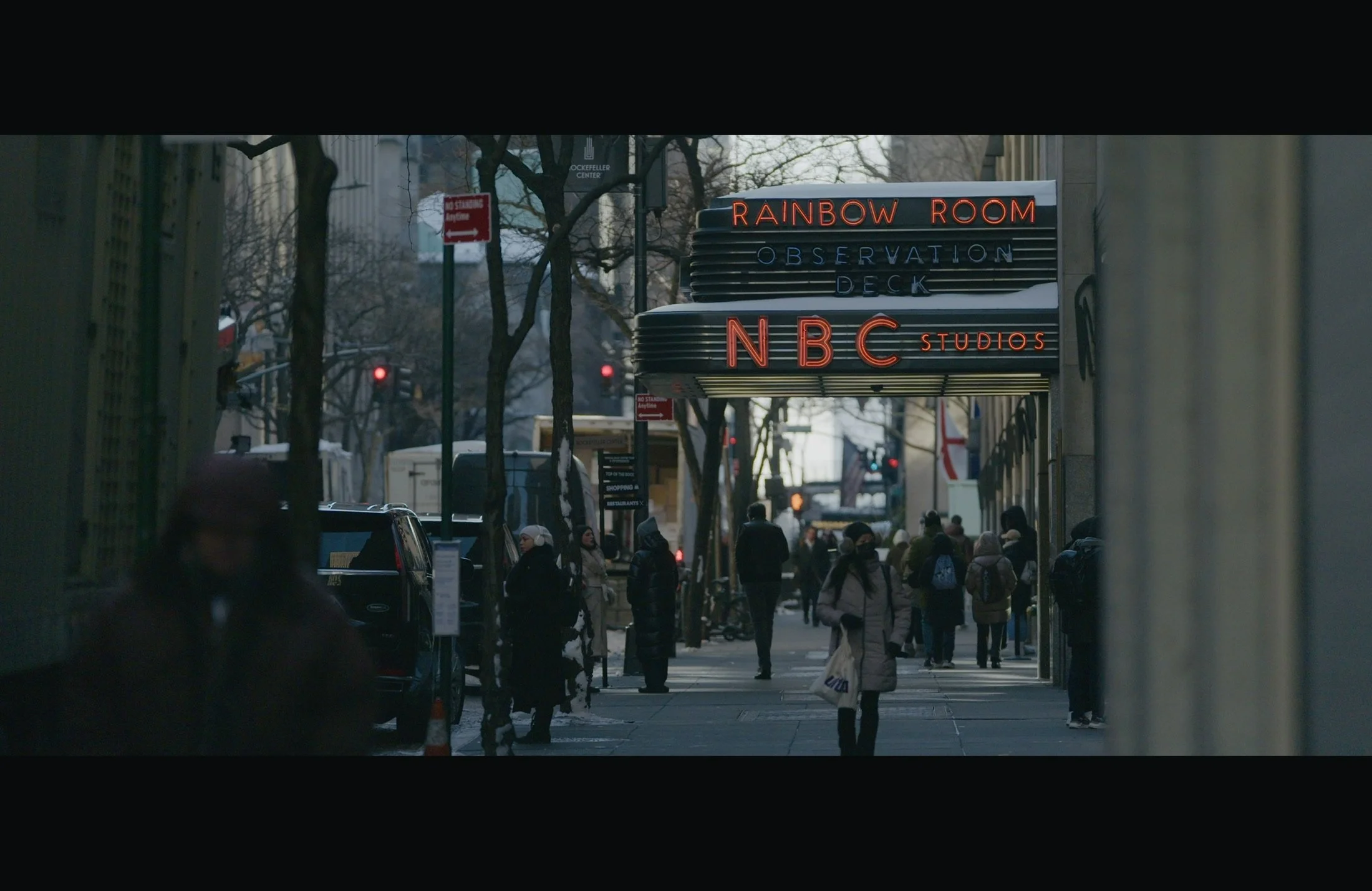 City street scene with pedestrians walking, trees bare of leaves, and a marquee sign reading "Rainbow Room Observation Deck NBC Studios"