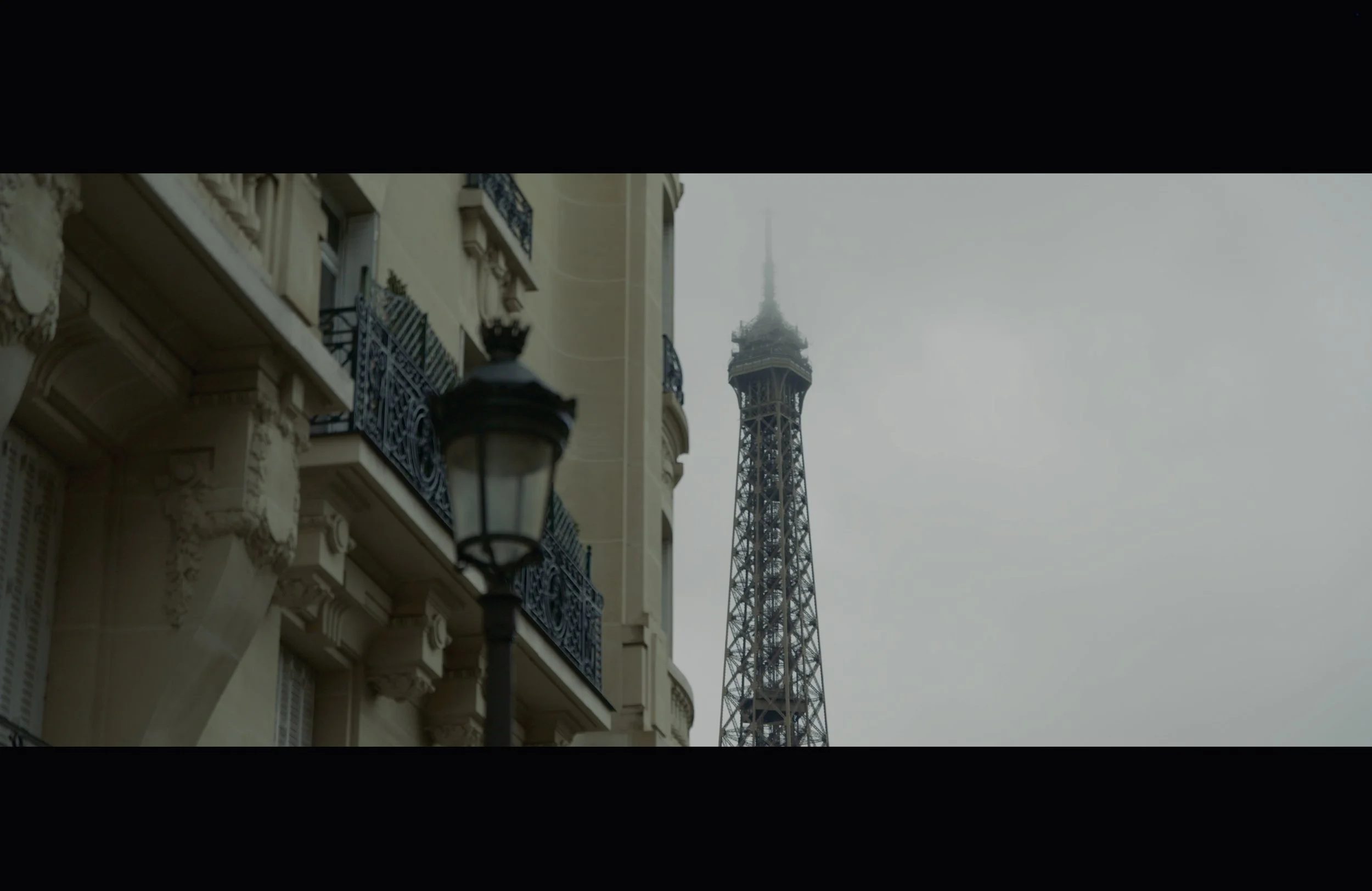 Parisian street with ornate buildings and a streetlamp in the foreground, the Eiffel Tower visible in the background on a cloudy day.