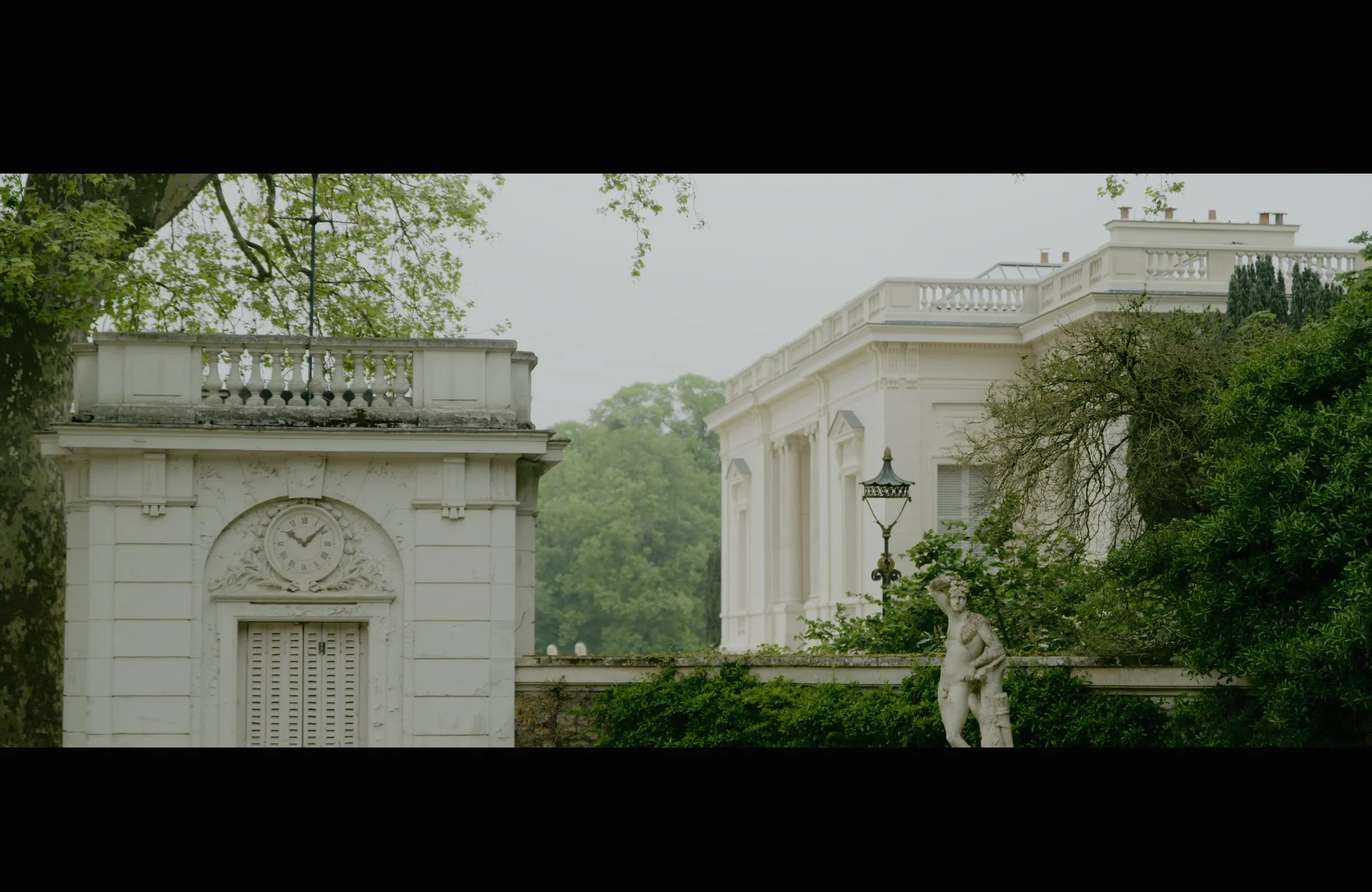 A white classical building with ornate architectural details, surrounded by lush greenery and trees, with a clock on the facade.