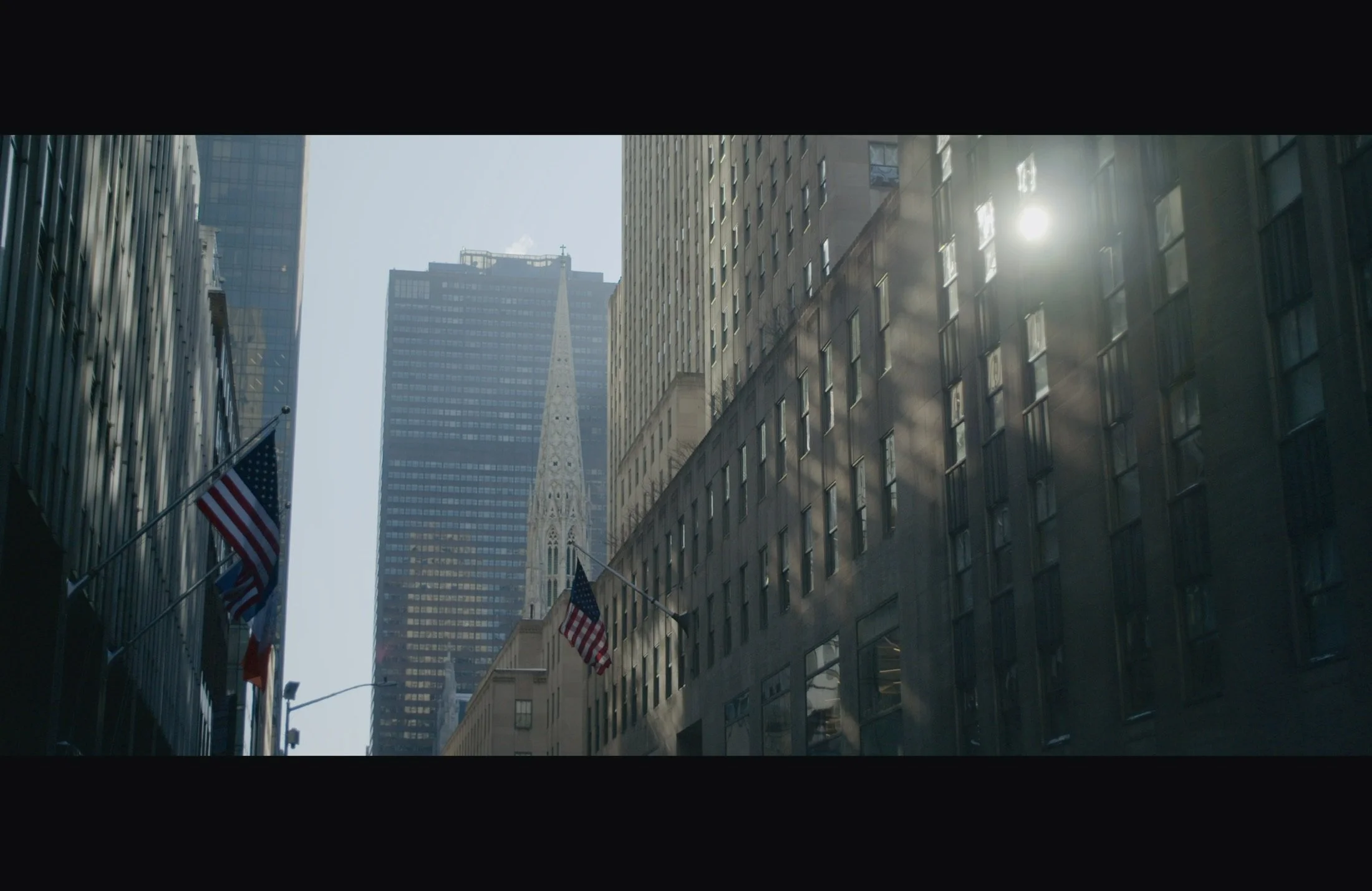 Tall skyscrapers in a city with American flags hanging from buildings, sunlight reflecting off windows, and a church spire rising between the buildings.