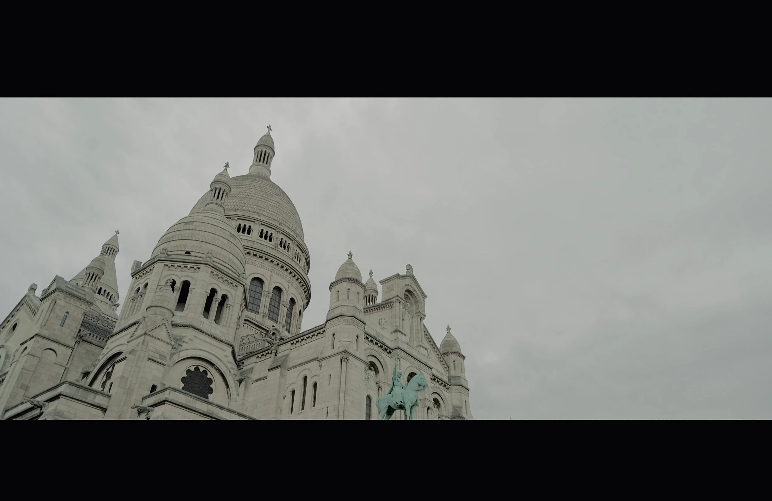 View of a historic European-style church or cathedral with white stone architecture, multiple spires, and a statue of a mounted figure at the front, under an overcast sky.