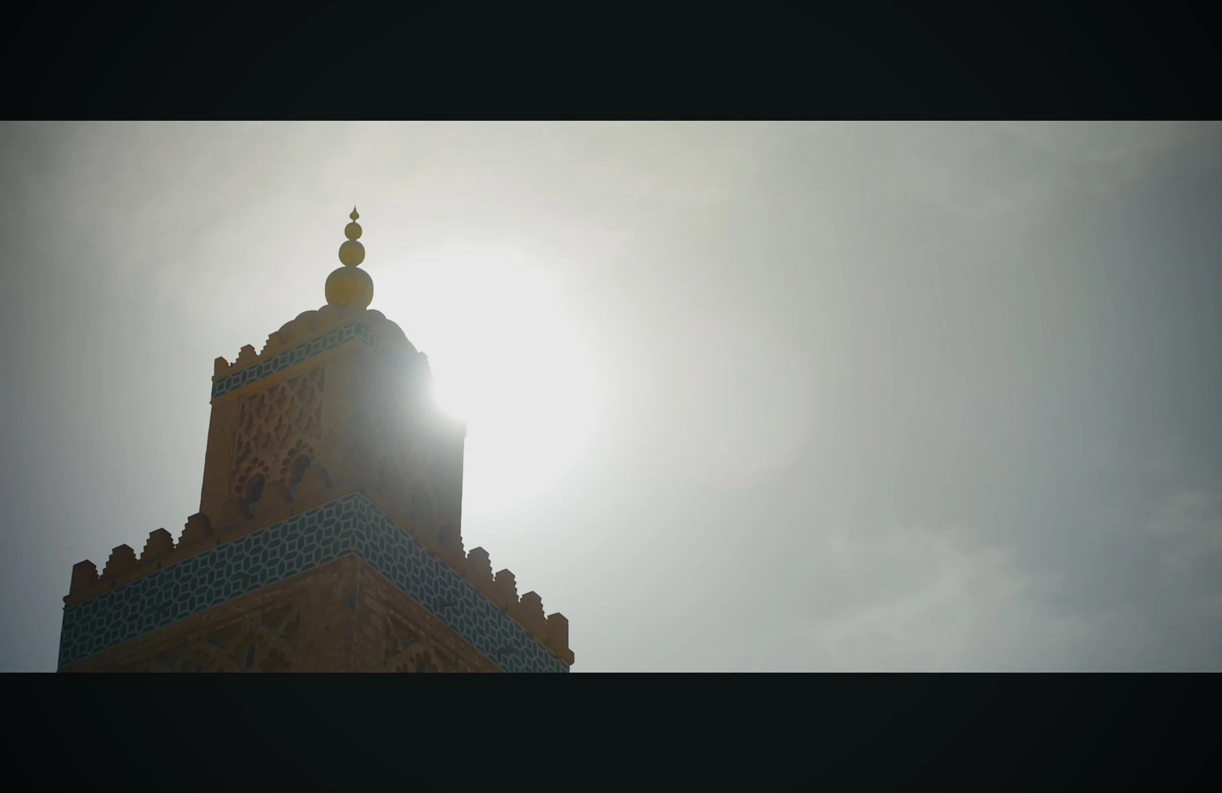 A tall, ornate tower with Islamic architectural design, topped with a decorative spire, is seen against the bright sun.
