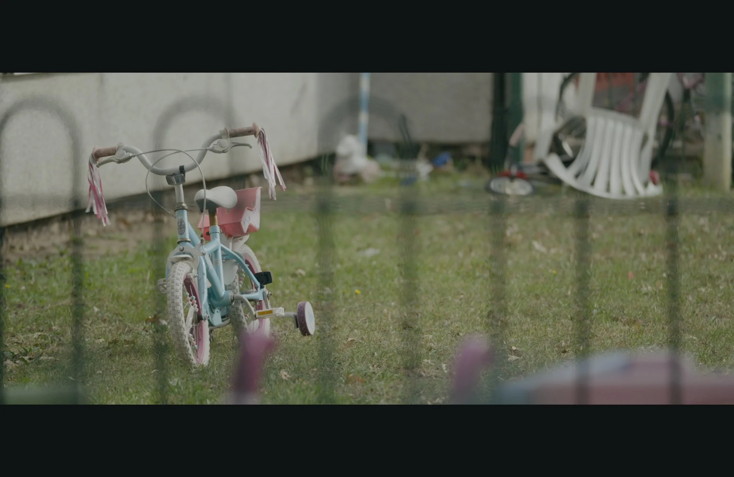 A pink and white children's bicycle with training wheels parked on grass near a concrete wall. A white outdoor chair and some scattered objects are in the background.