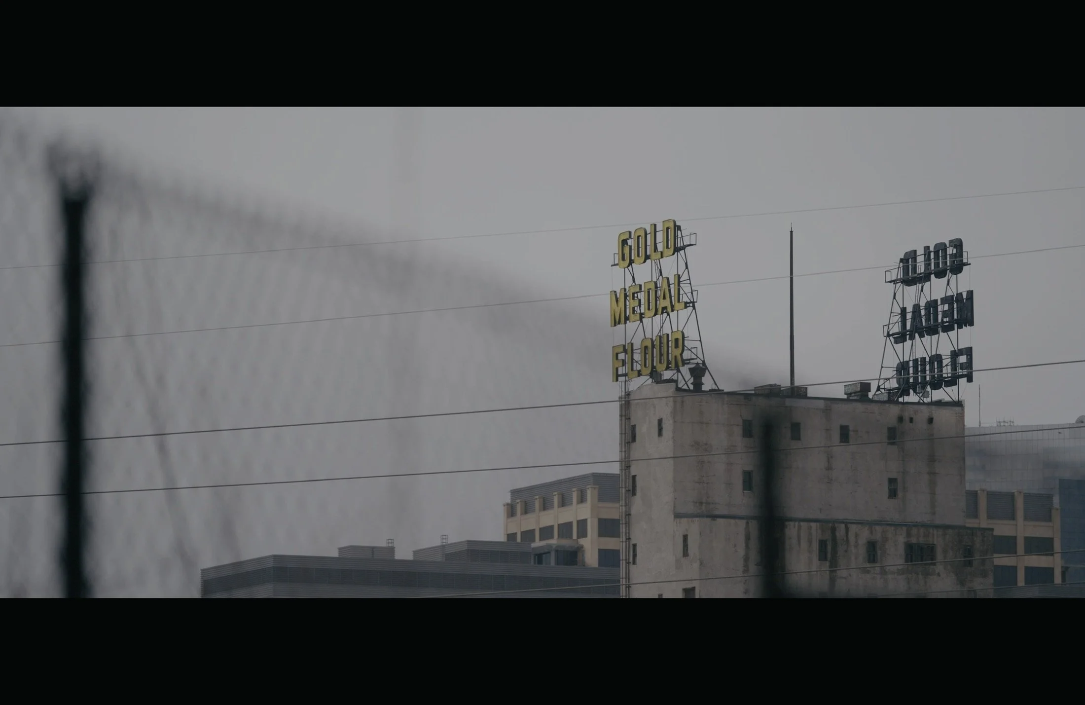 A building with illuminated signs, including one that says 'GOLD MEDAL FLOUR' and another that says 'HAGEN' on a rooftop. The sky is overcast and there are power lines in the foreground.