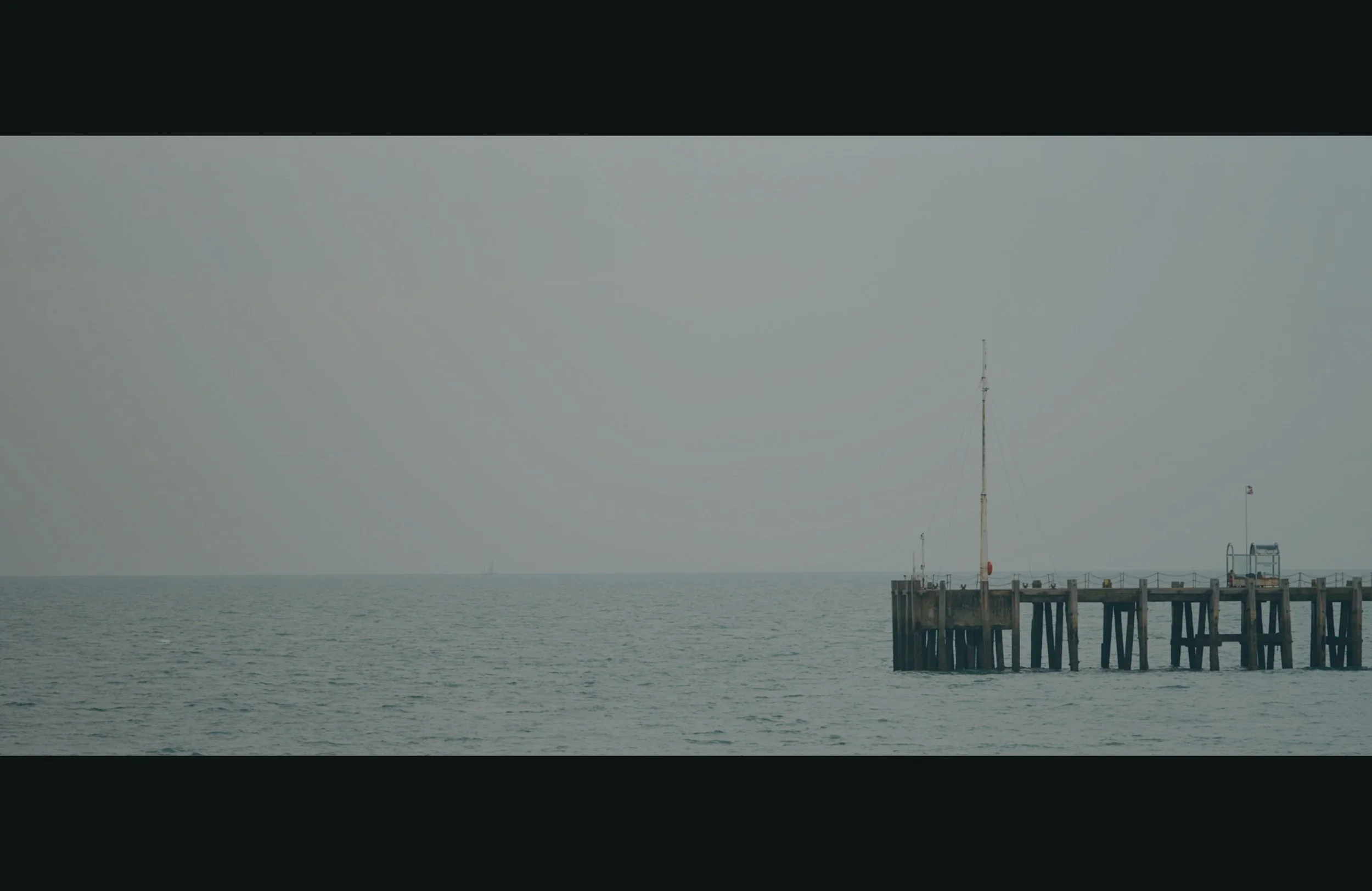 A small pier with a boat docked and a tall radio tower in the distance over calm water and an overcast sky.