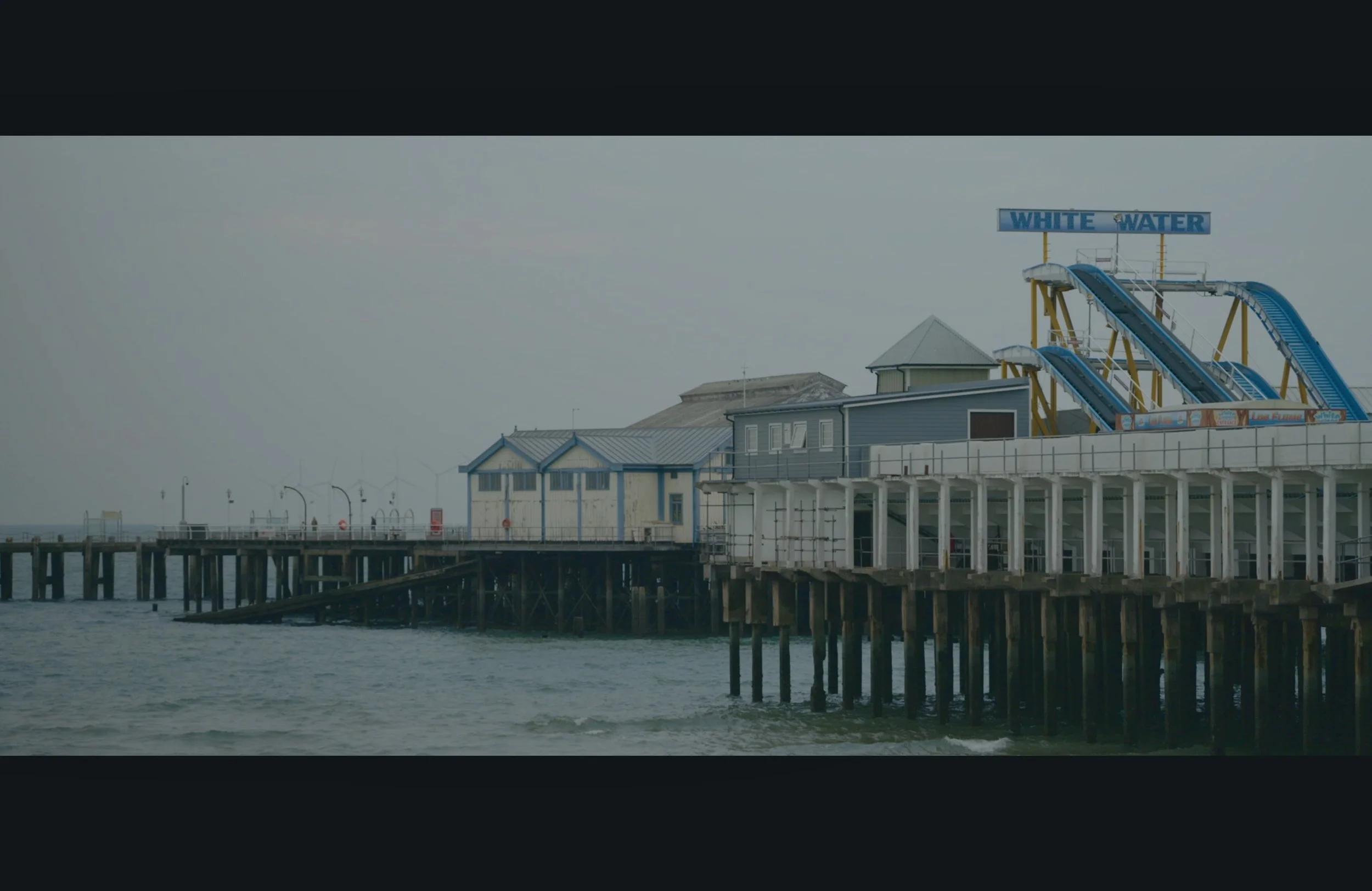 A seaside pier with buildings and a water slide structure labeled 'White Water' extending above the buildings. The pier is supported by wooden pilings over the water, with a cloudy sky in the background.