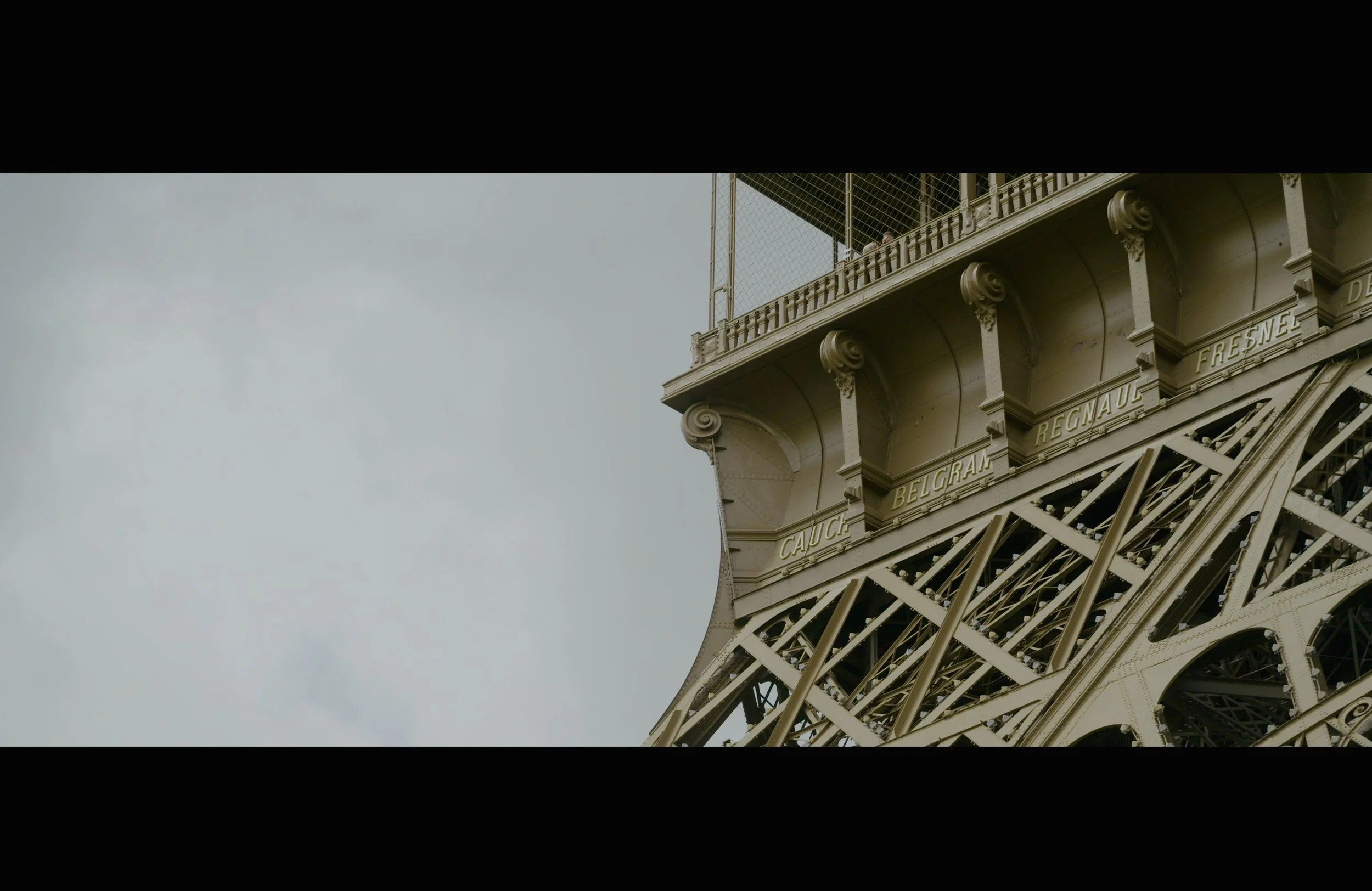 Close-up of the Eiffel Tower's iron lattice structure with names of regions such as 'CAUC', 'BELGRA', 'REGNAUE', and 'FRESNE' inscribed on it, and a section of the sky visible in the background.