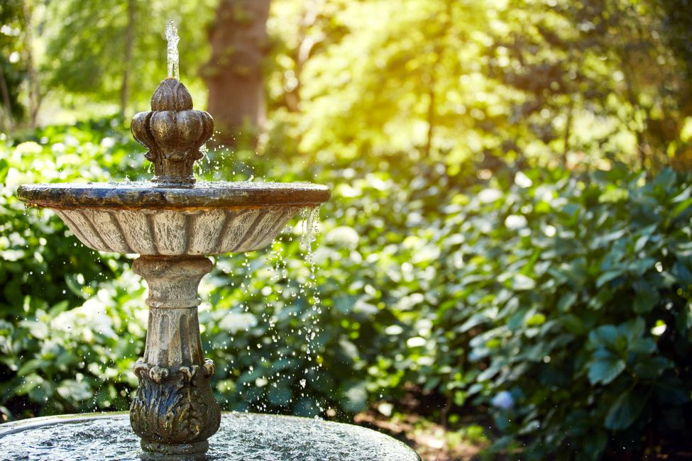 A stone fountain with water flowing in a garden or park, surrounded by green foliage and trees, with sunlight filtering through leaves.