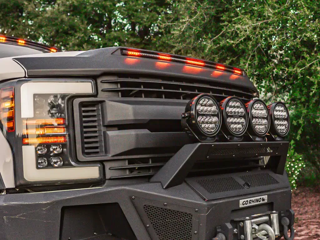 Close-up of the front of a rugged off-road vehicle with multiple large lights mounted on the grille, featuring a black and white body, against a background of green trees.