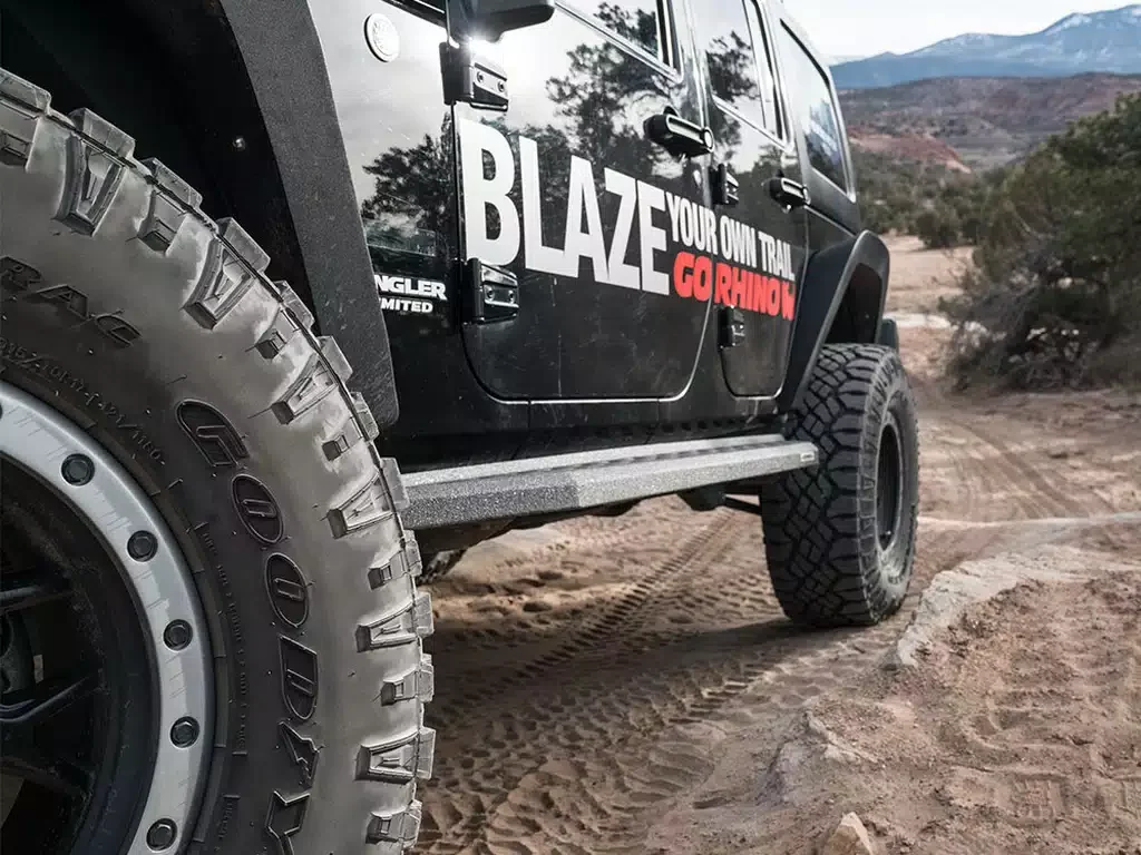 Close-up of a black Jeep Wrangler with off-road tires on a dirt trail in a desert landscape, showing the side door with branding that reads 'Blaze Your Own Trail GORhino'.