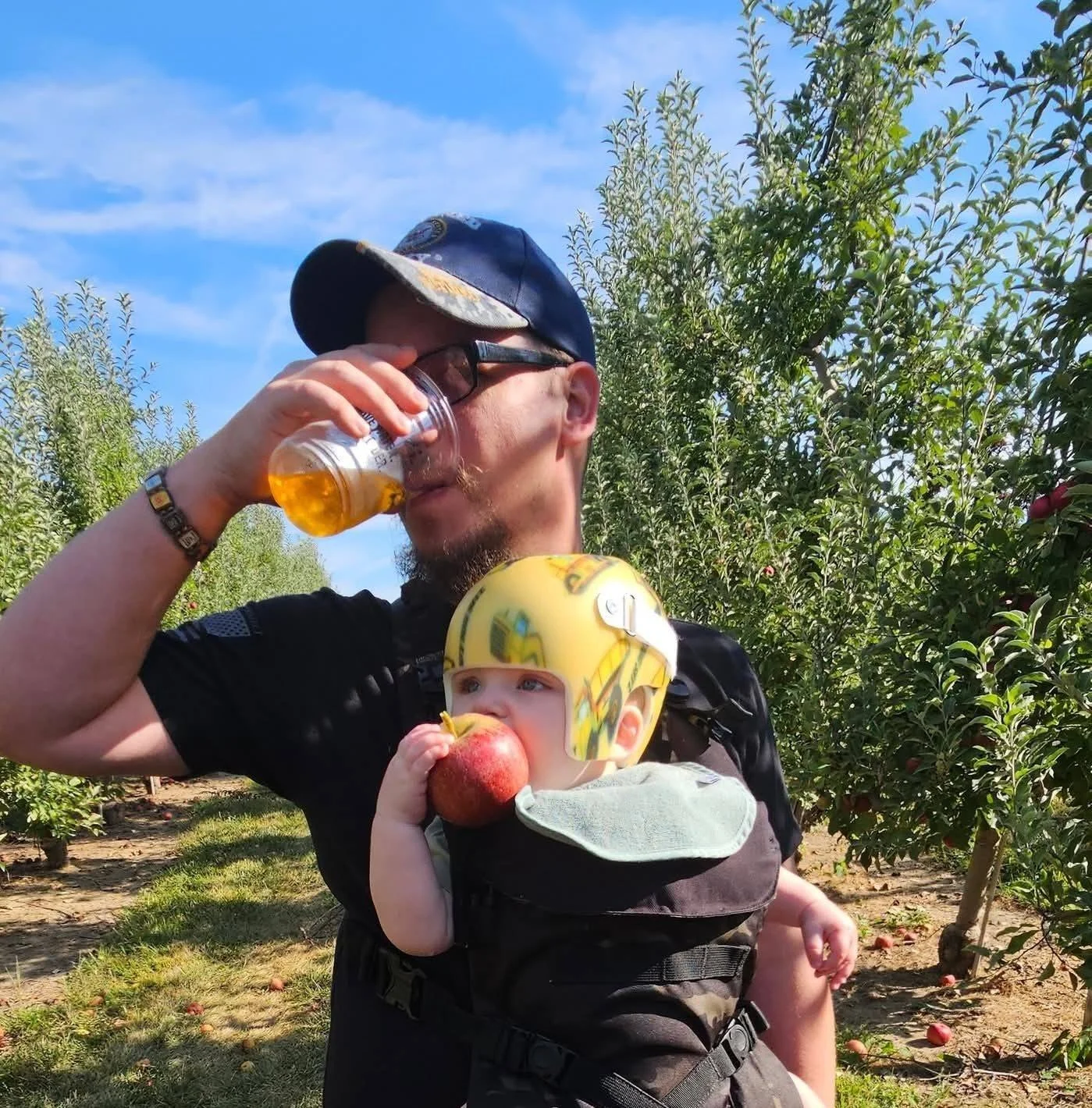 Man wearing a cap and sunglasses drinking from a cup, carrying a child in a baby carrier. The child is wearing a helmet and holding an apple. They are outdoors in an orchard with trees and blue sky.