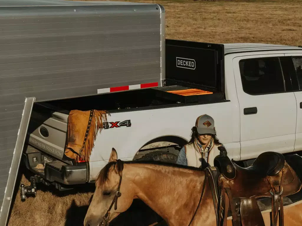 A young girl in a cowboy hat and vest standing next to a brown horse with a saddle in a field, with a white truck and trailer in the background.