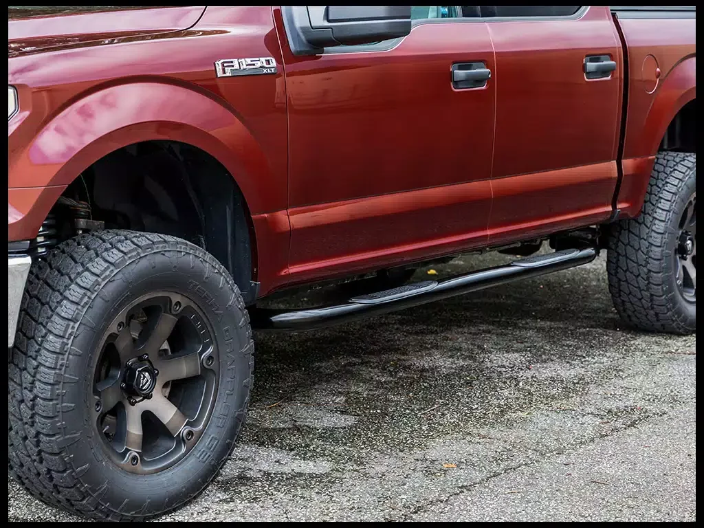 Red Ford F-150 XLT pickup truck with off-road tires and black side step bar, parked on a wet asphalt surface.