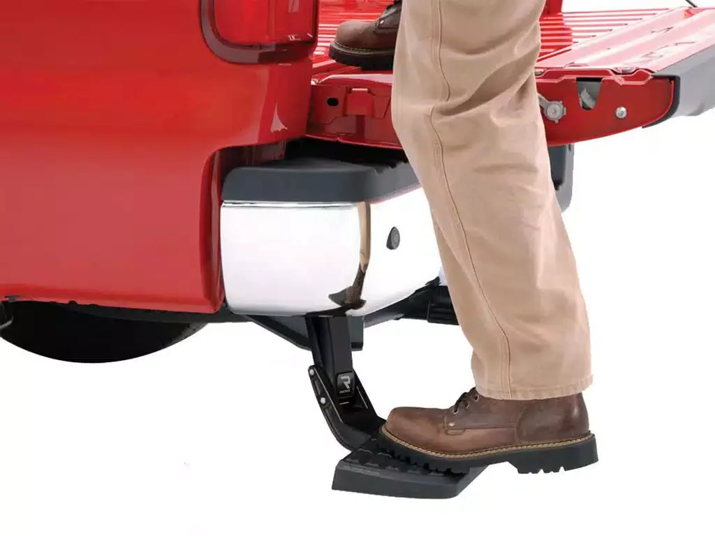 Close-up of a person’s feet on motorcycle foot pegs, wearing brown boots and beige pants, motorcycle is red, and the background is white.