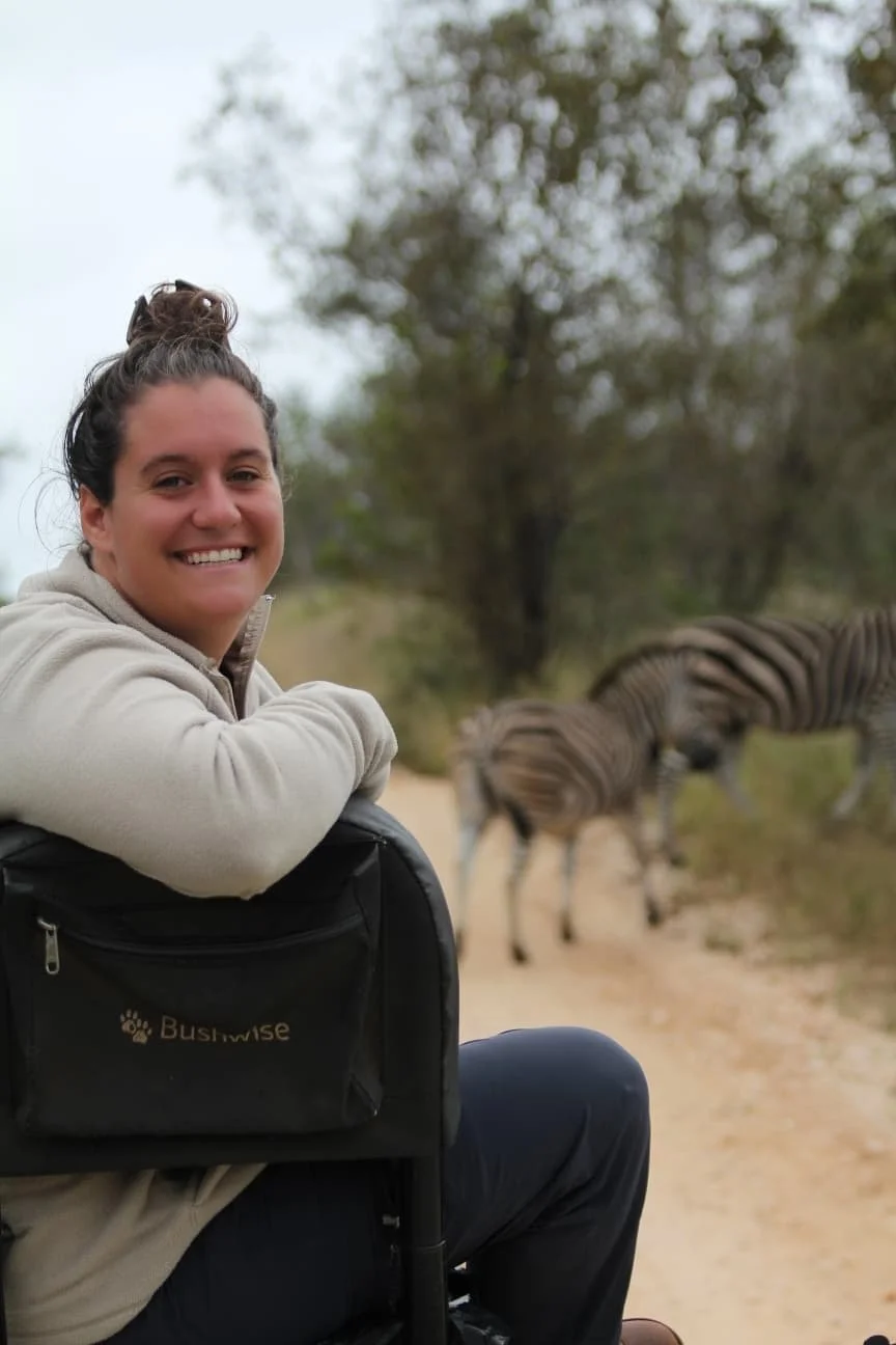 Woman smiling in a safari vehicle with zebras grazing in the background.