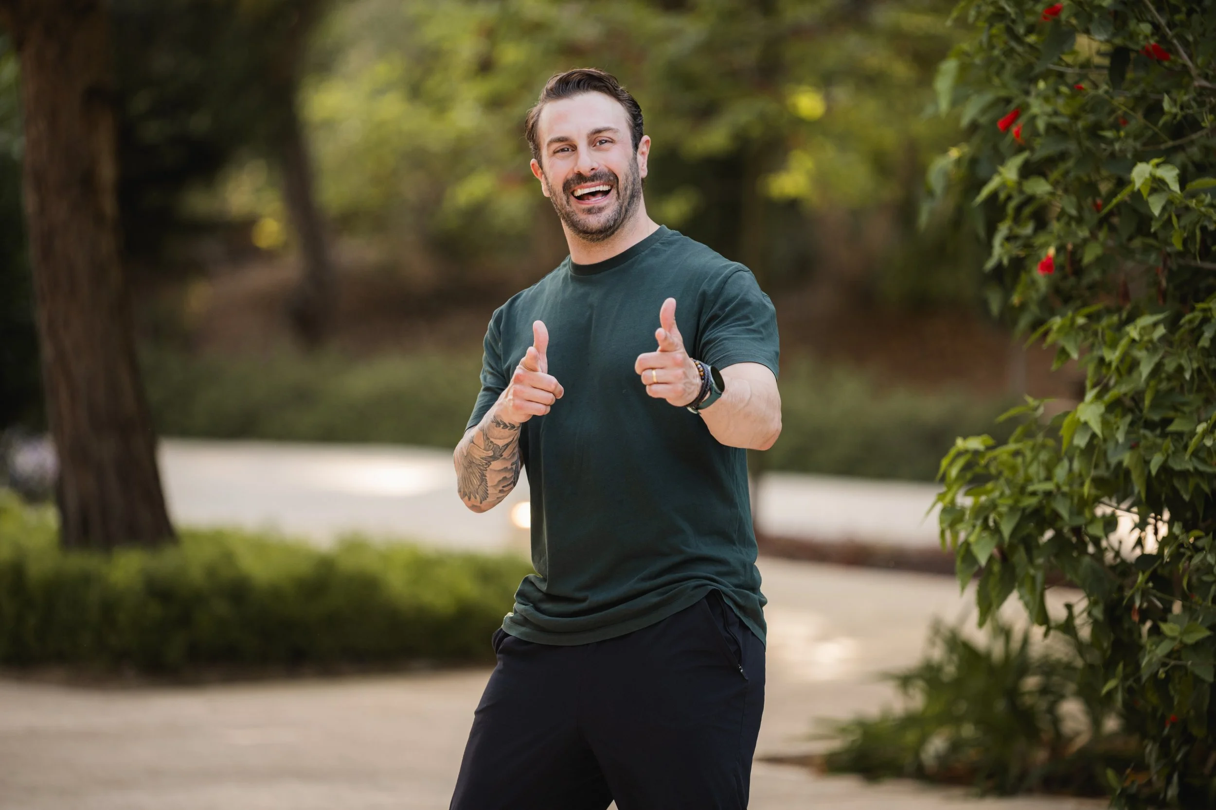 A smiling man with tattoos on his right arm is pointing at the camera with both hands while walking outdoors in a park or garden with trees and bushes. Happy