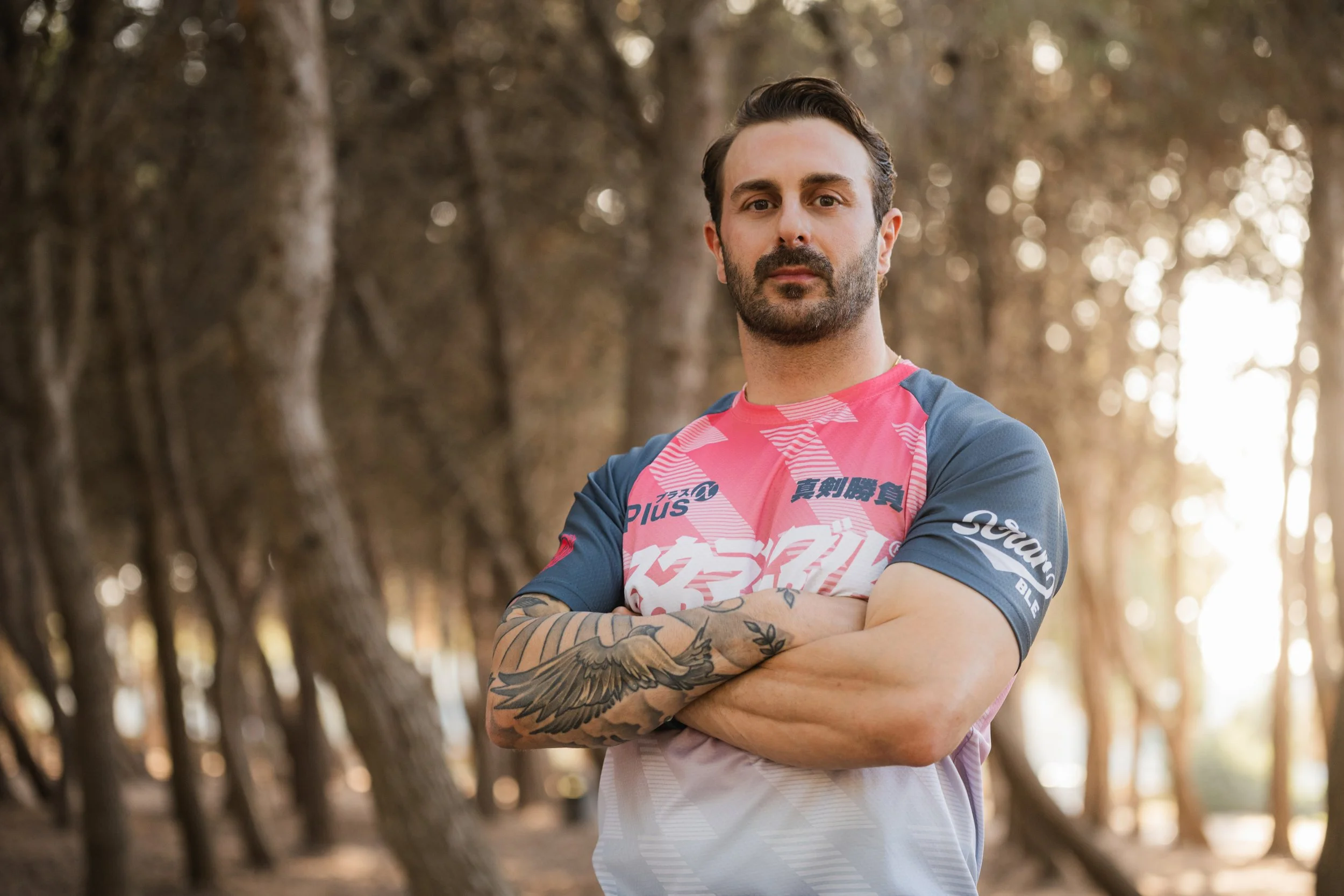 A man with dark hair and a beard standing outdoors with arms crossed, wearing a pink and blue athletic shirt, and surrounded by trees with sunlight in the background. looks athletic.