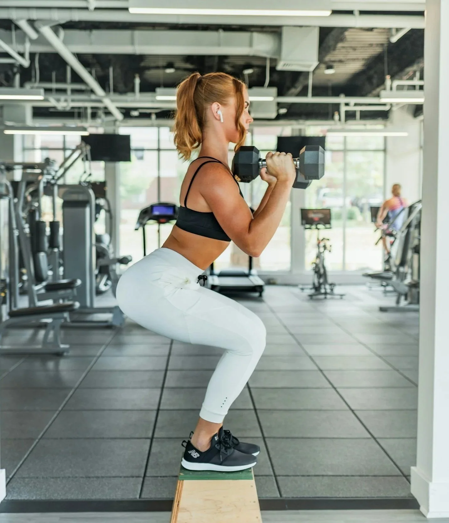 A woman in athletic wear performing a squat exercise with dumbbells on her shoulders at the gym, standing on a small wooden platform looking lean and toned.