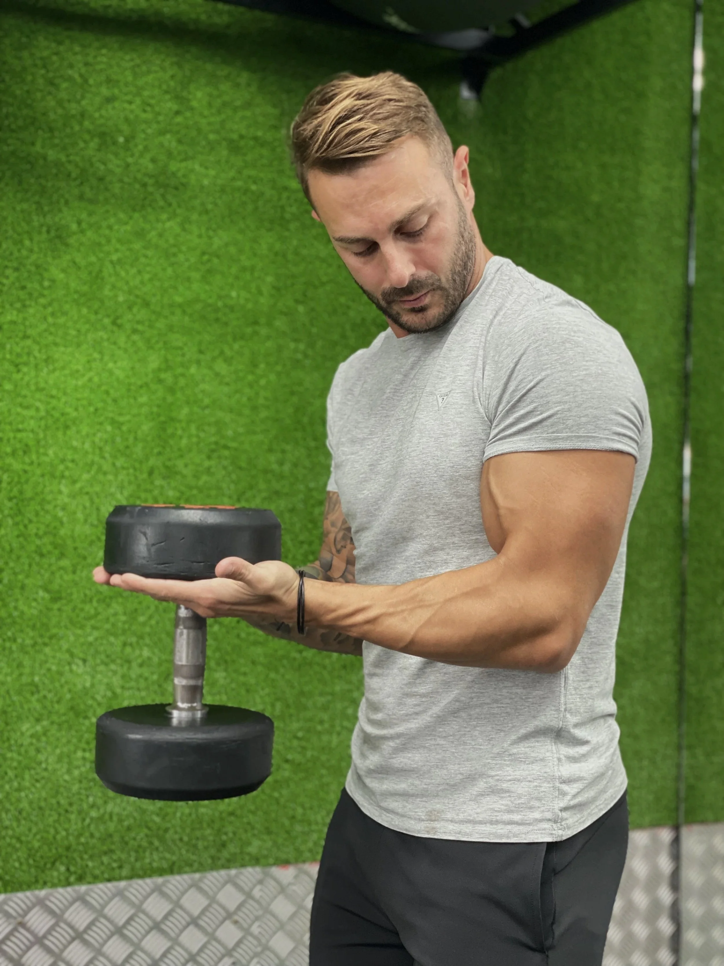 A man with brown hair and a beard, wearing a light gray T-shirt and black pants, is holding a large weight plate in a gym with green padding on the walls with big biceps.