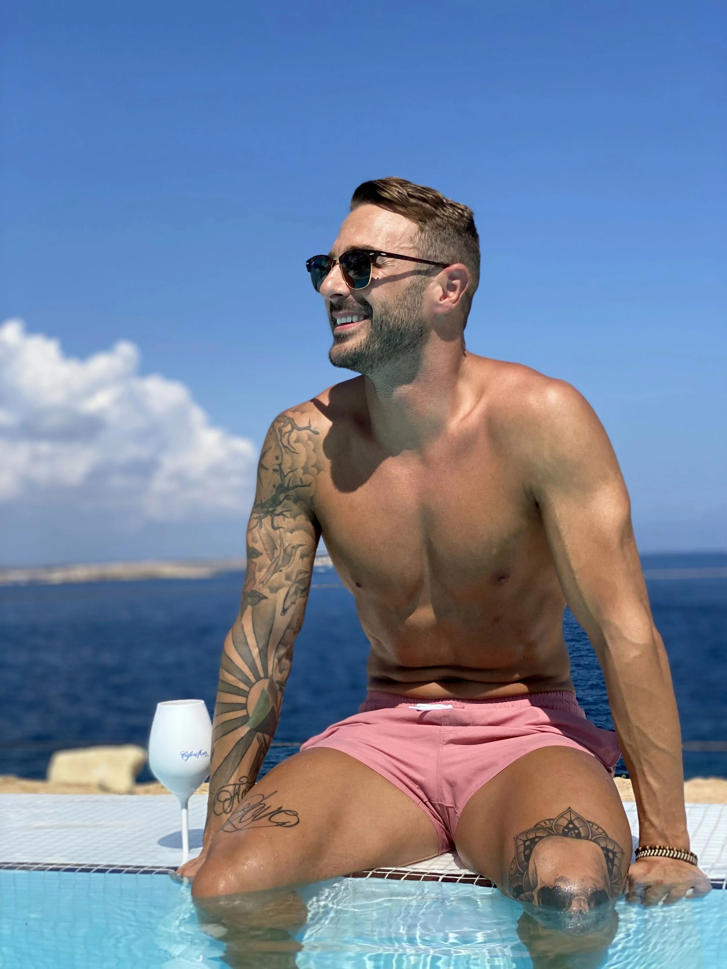 A man with sunglasses and tattoos sitting at the edge of a swimming pool with the ocean in the background, wearing pink swim trunks with a great looking beach body.