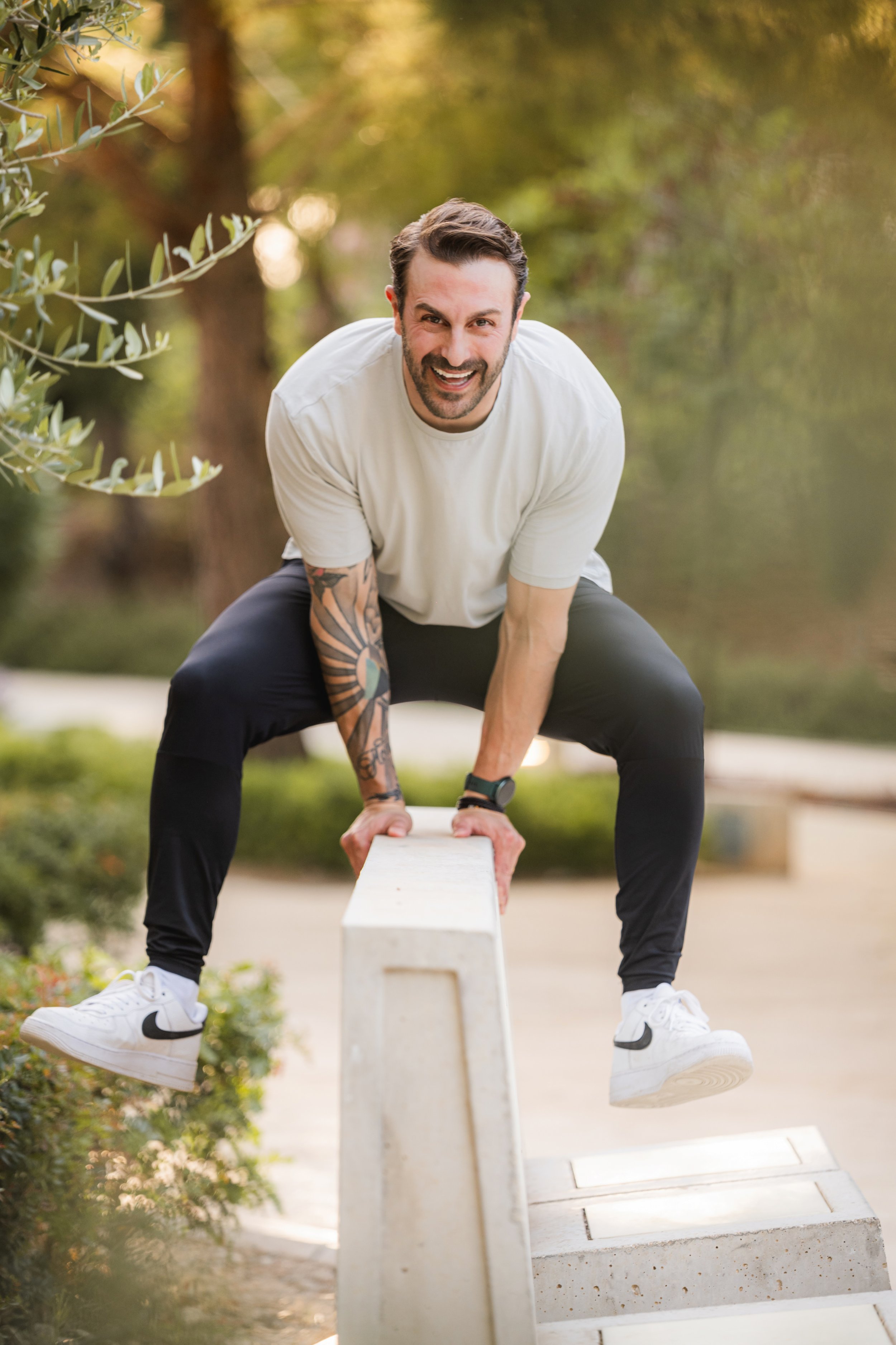 A man with a beard and tattoos on his left arm is crouching on a balance beam, appearing to be about to fall forward. He is wearing a white t-shirt, black pants, and white Nike sneakers with black swooshes. The background is an outdoor park with green trees and bushes, captured in warm lighting.
