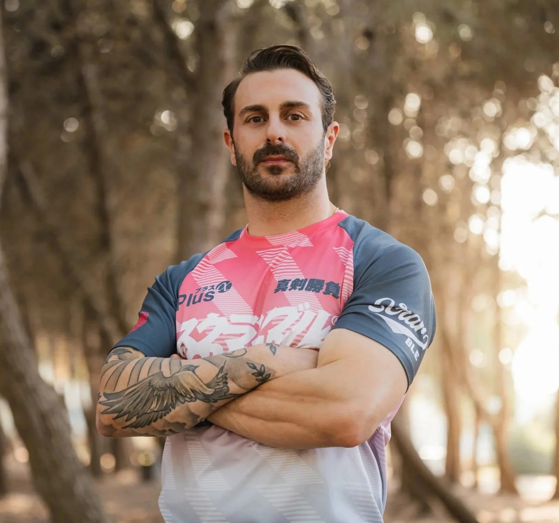 A man with dark hair, a beard, and tattoos on his arms standing outdoors with his arms crossed, wearing a colorful sports jersey, in a forest setting with blurry trees and sunlight in the background.