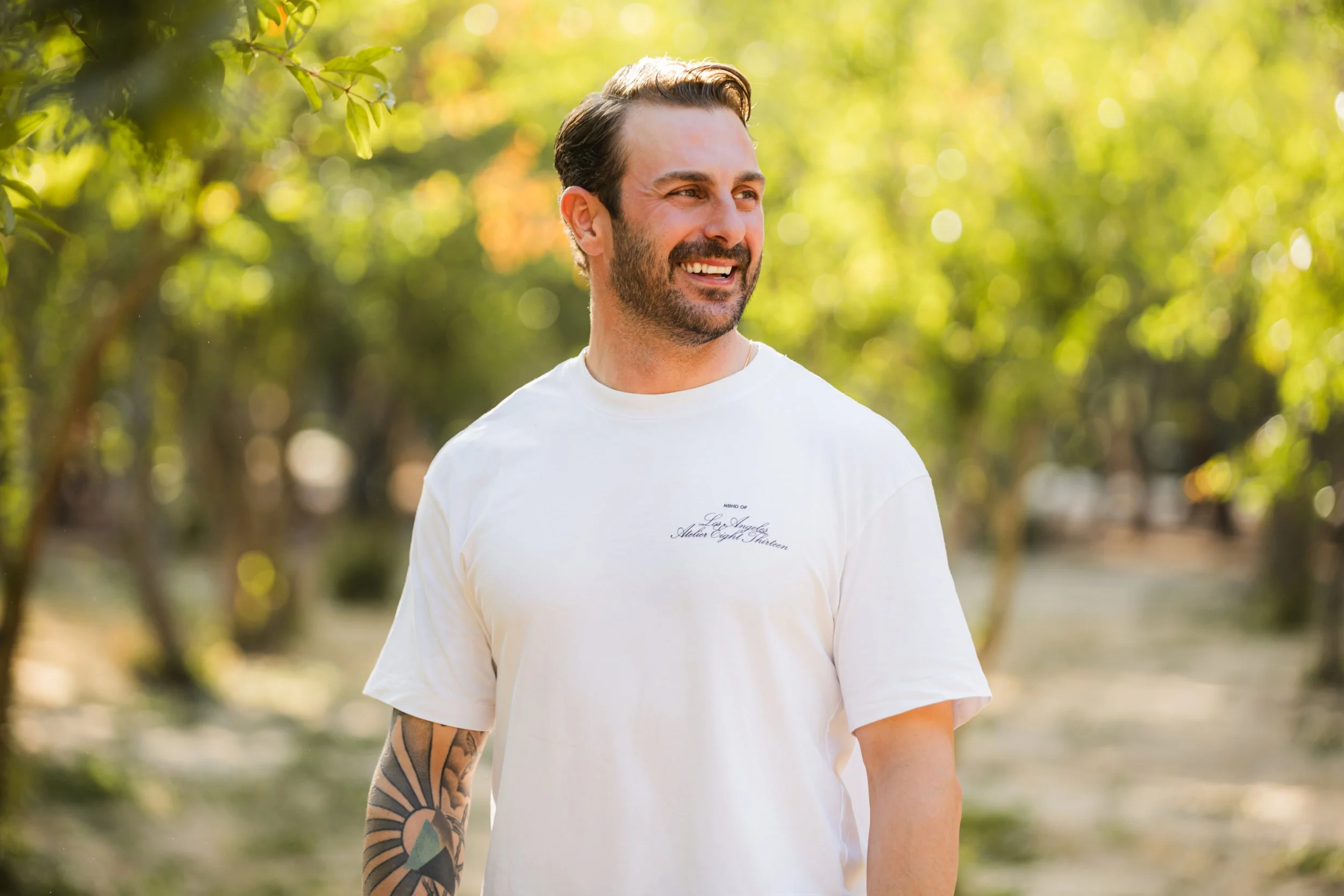A smiling man with dark hair and beard wearing a white t-shirt with text on it, walking outdoors in a park with trees and green foliage in the background.