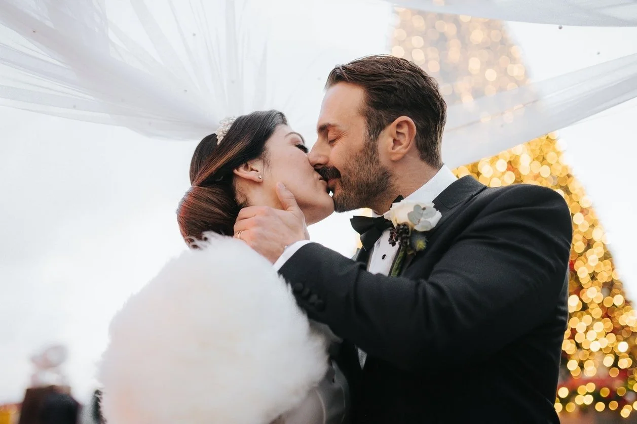 A couple dressed in wedding attire sharing a kiss at their wedding, with festive Christmas lights in the background.