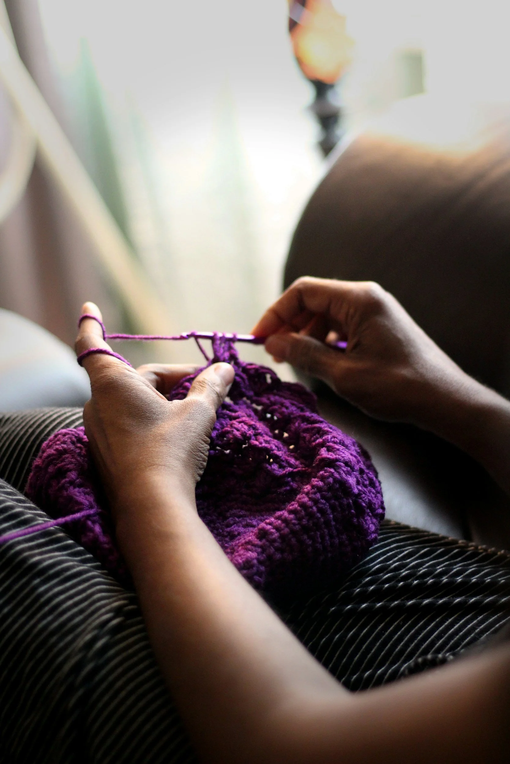 Person knitting with purple yarn, sitting indoors with sunlight in the background