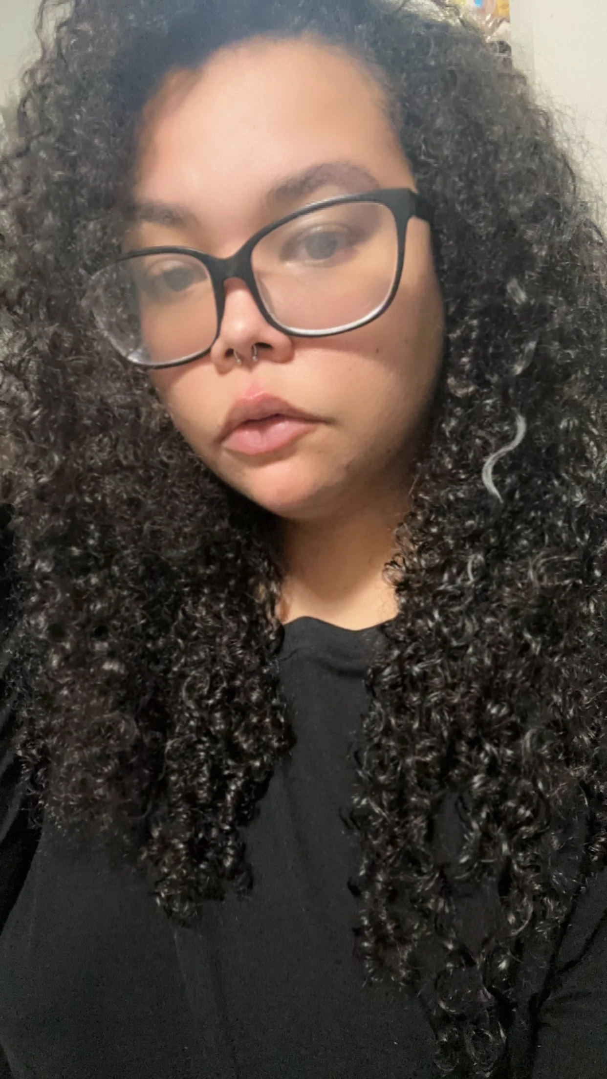 A woman with curly dark hair wearing glasses and a septum piercing, dressed in a black top.