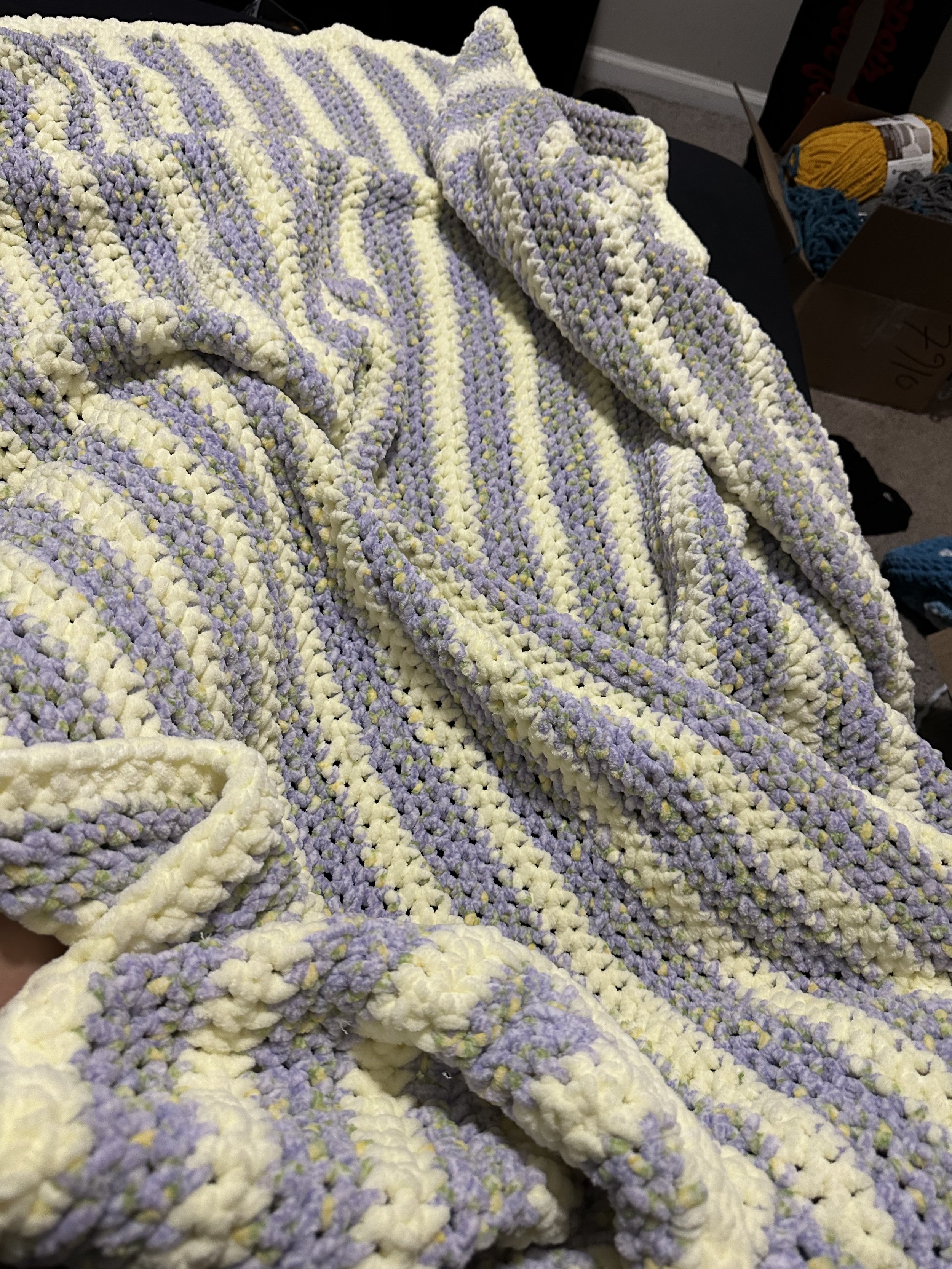 Close-up of a crocheted blanket in pastel yellow and purple colors with a striped pattern, draped over a surface with yarn balls and other crocheted items in the background.