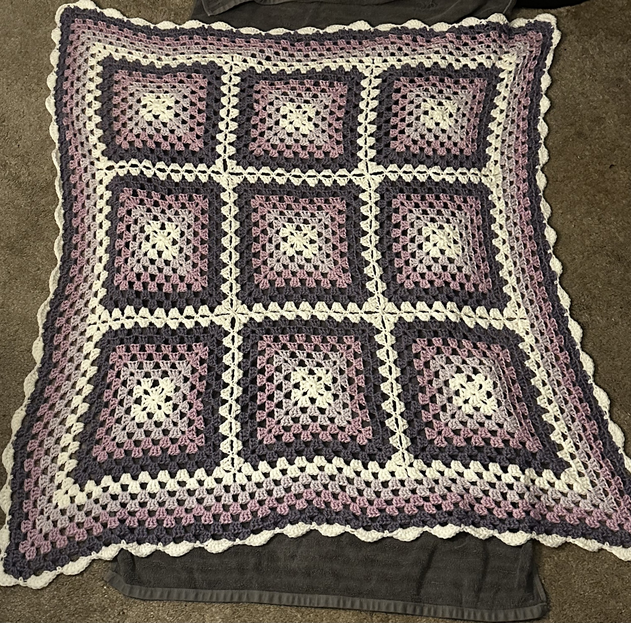 A crocheted blanket with a geometric pattern of squares in purple, white, and pink, bordered by a white edge, laid out on a dark surface.