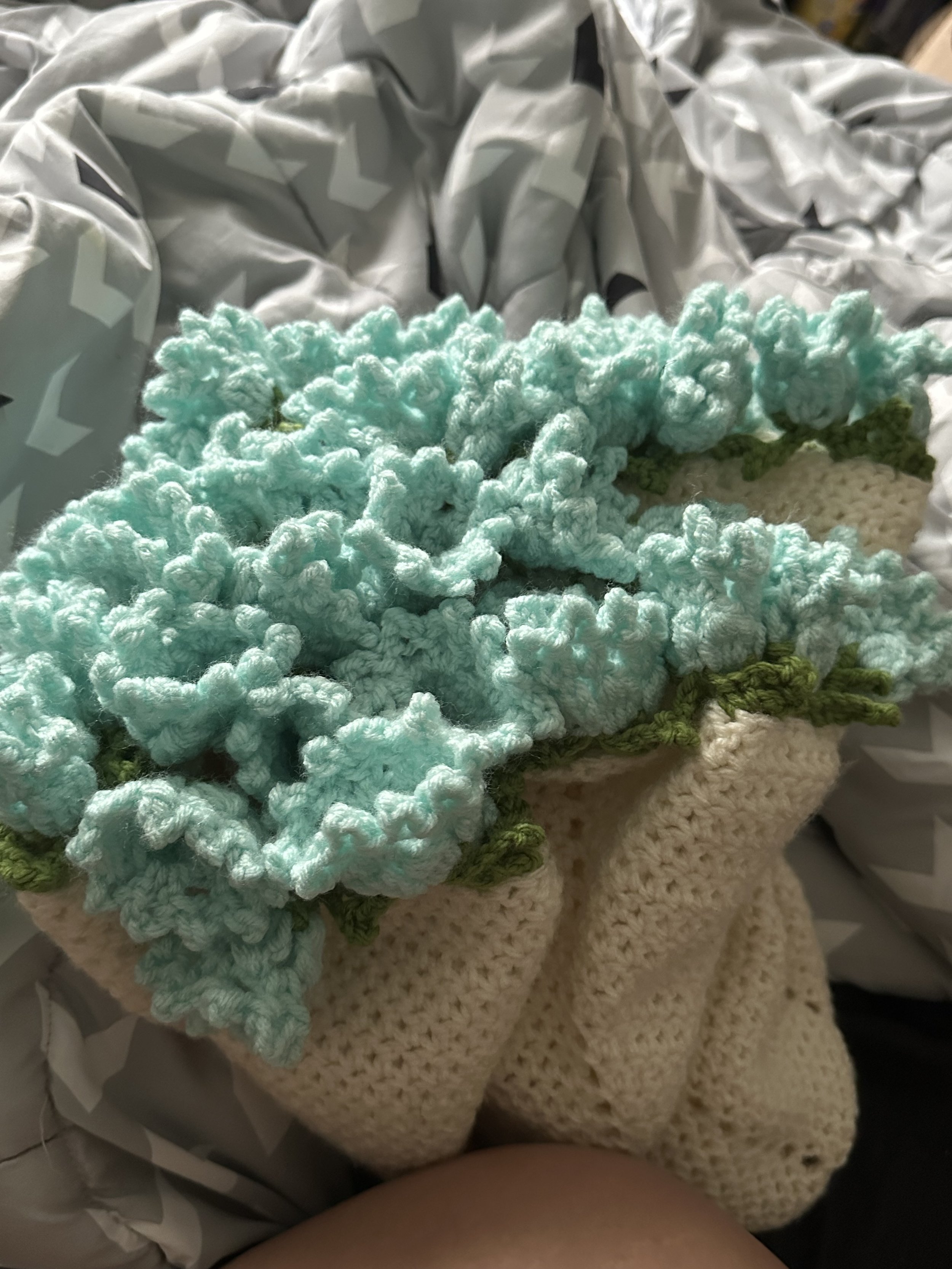 A close-up of a crocheted flower with light blue petals and green leaves, resting on a beige crocheted surface.