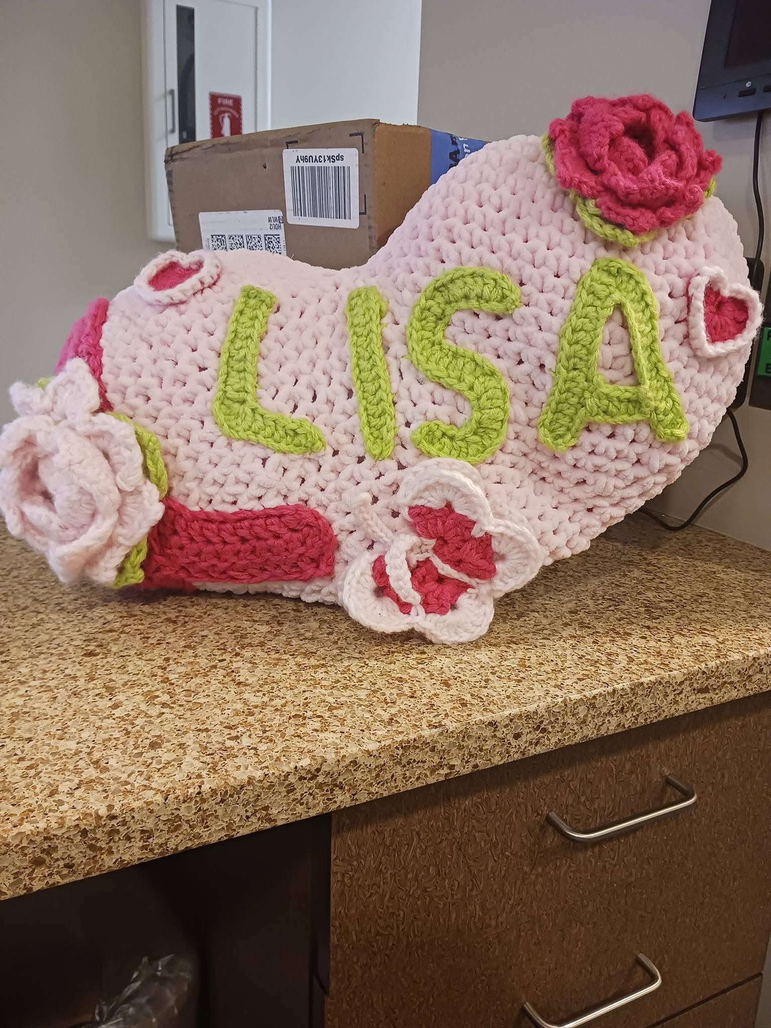 A knitted or crocheted item resembling a baby blanket or decoration with the name 'LISA' in green lettering. It has pink, red, and white floral accents, placed on a speckled beige countertop. Behind it is a cardboard box and a fire extinguisher cabin