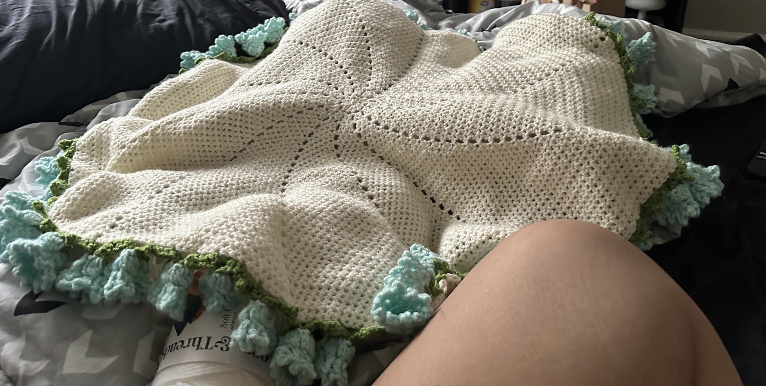 A crocheted blanket with a cream-colored body and a border made of light blue and green yarn, resting on a bed with bedsheets.