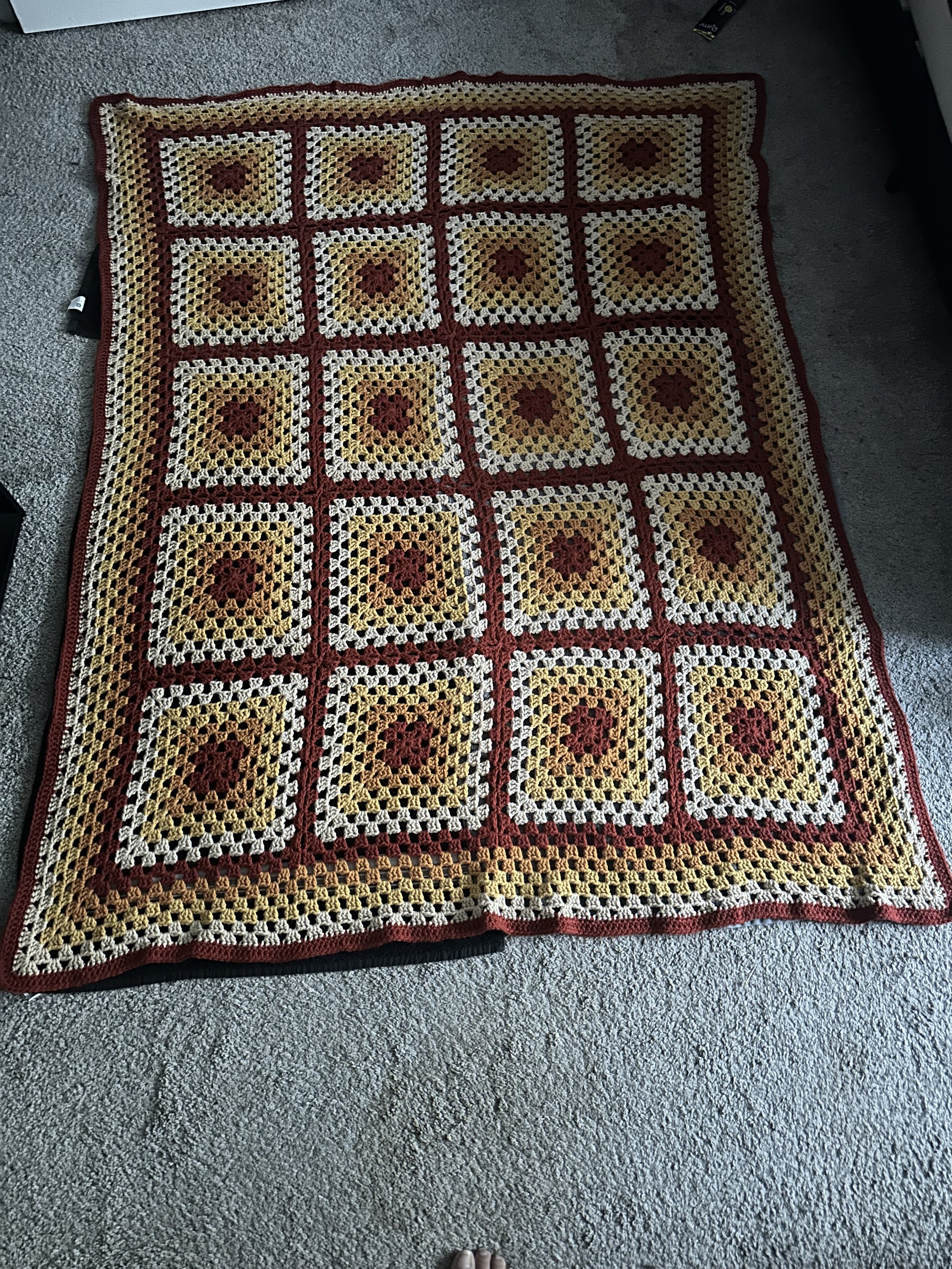 A colorful crocheted blanket with a granny square pattern, laid on a gray carpeted floor.