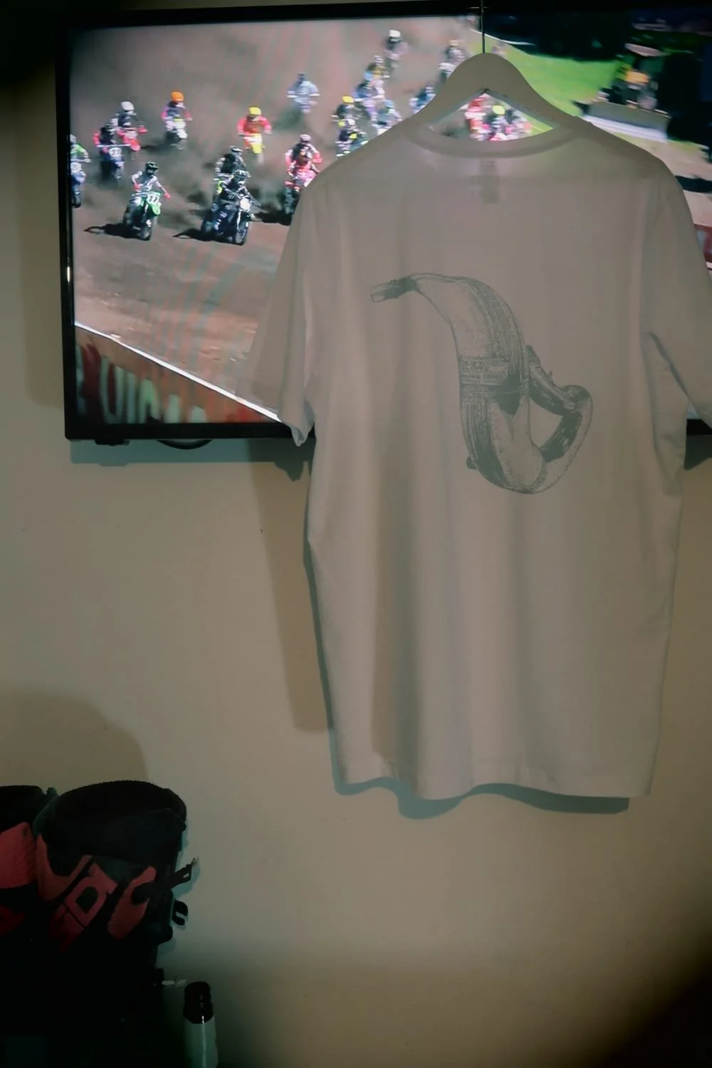 White moto t shirt with 2 stroke expansion pipe design hangs in front of TV showing motocross racing