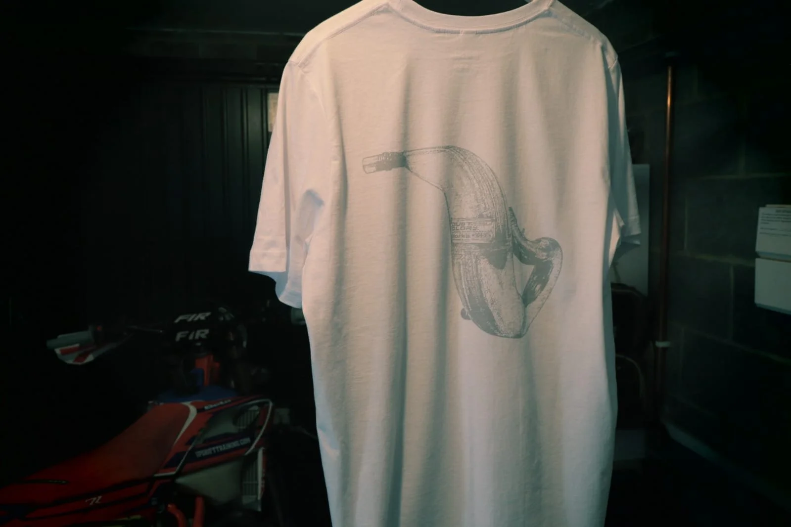 White t shirt with 2 stroke exhaust graphic design hangs in garage with enduro bike in the background