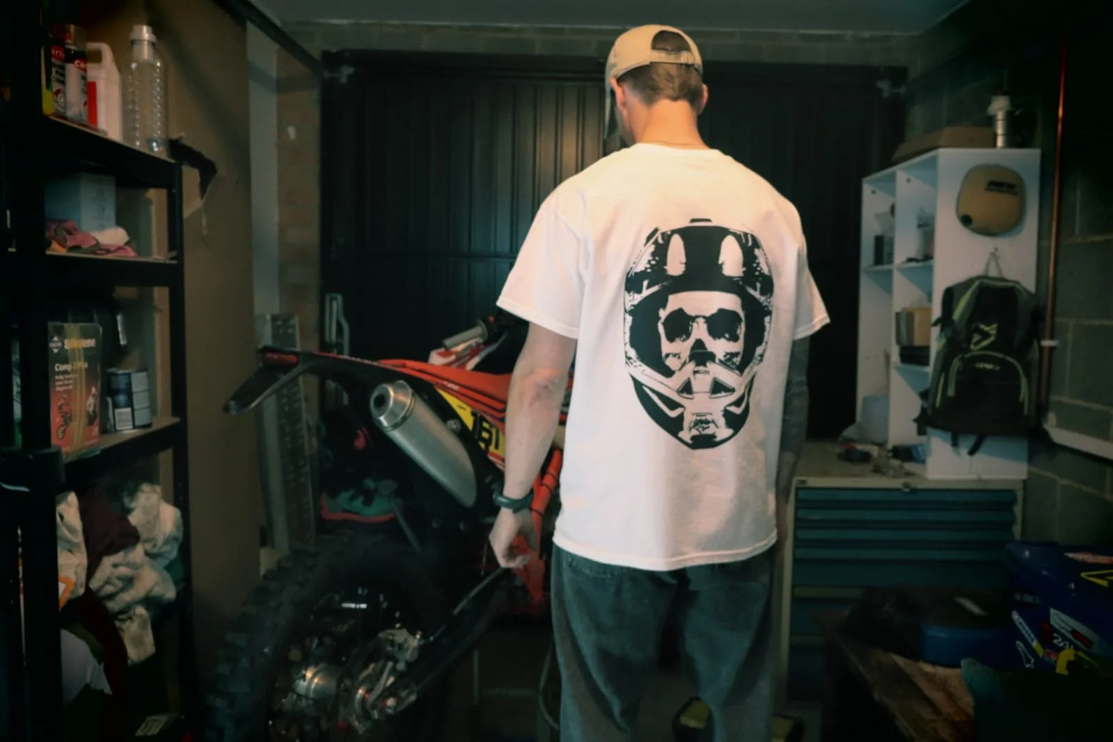 Man wearing white moto skull design graphic t shirt, staring at enduro motocross bike in garage
