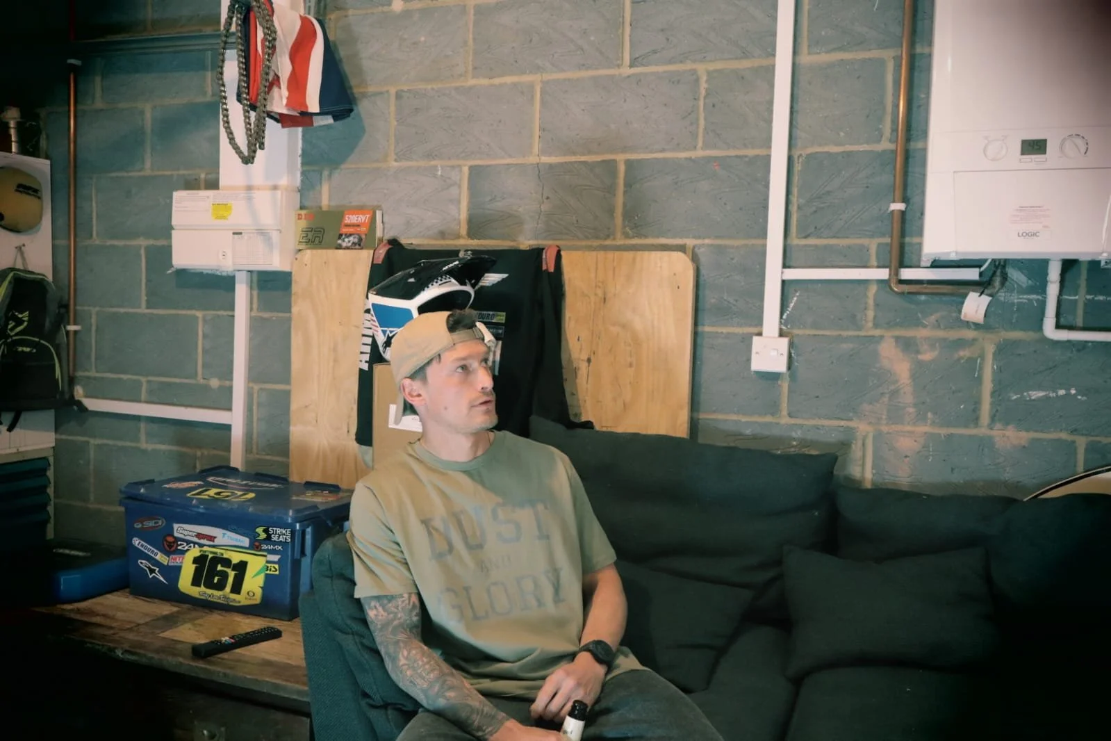 Man sitting in garage wearing a khaki Dust and Glory t shirt and drinking a beer