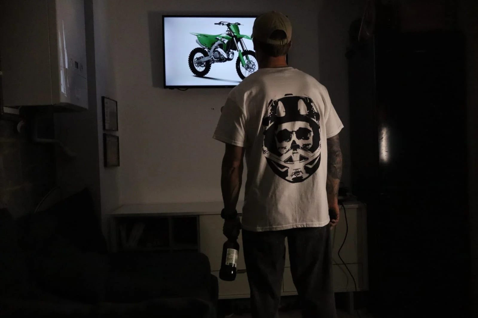 Man standing in garage staring at TV with Kawasaki KX 327 on the screen, holding beer, wearing white moto skull graphic design t shirt