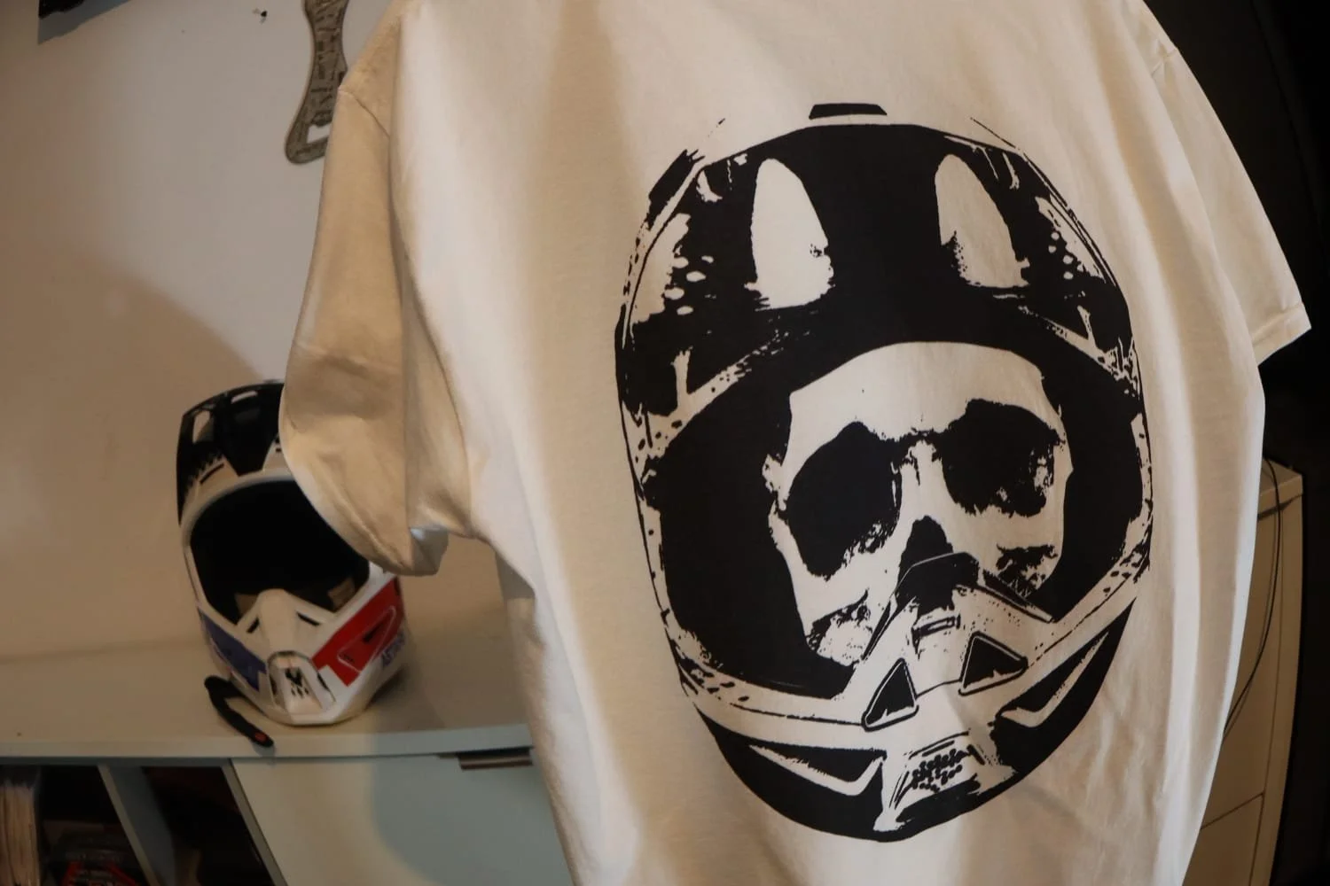 White moto skull design t shirt hangs in garage with motocross helmet in the background