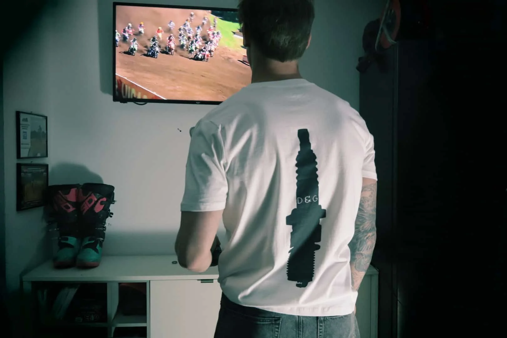 Image of model in garage with beer watching motocross on TV and wearing white moto t shirt with spark plug design on the back