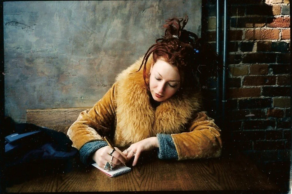 A woman with reddish dreadlocks writing in a small notepad at a wooden table, wearing a brown fur-lined coat, in an industrial-style setting with a gray wall and brick wall in the background.