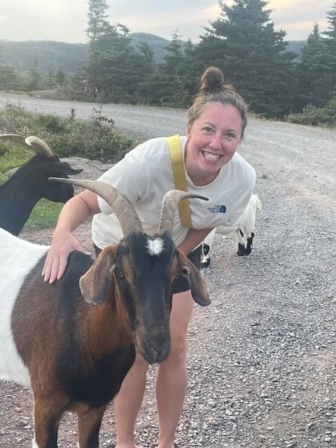 A young woman with a top knot hairdo smiling and petting a goat with large curved horns on a gravel path in a wooded outdoor area.