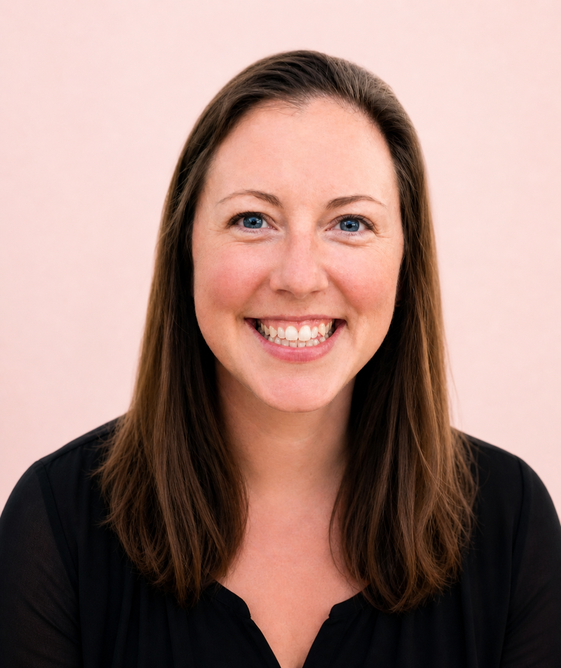 A woman with long brown hair, blue eyes, and a bright smile wearing a black top, standing against a light pink background.