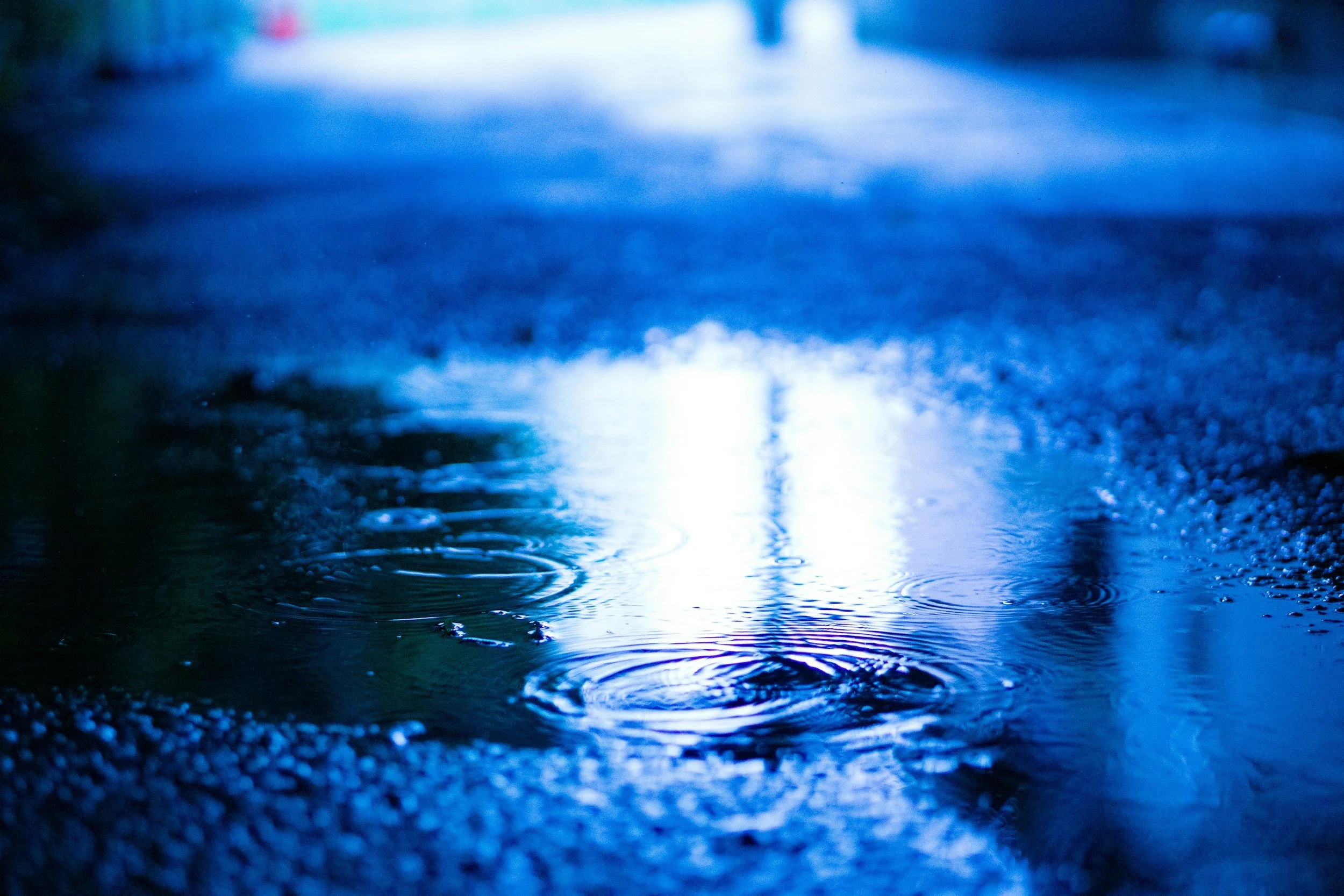 A close-up of a wet asphalt road at night with reflections of blue and white lights, and water droplets creating ripples.