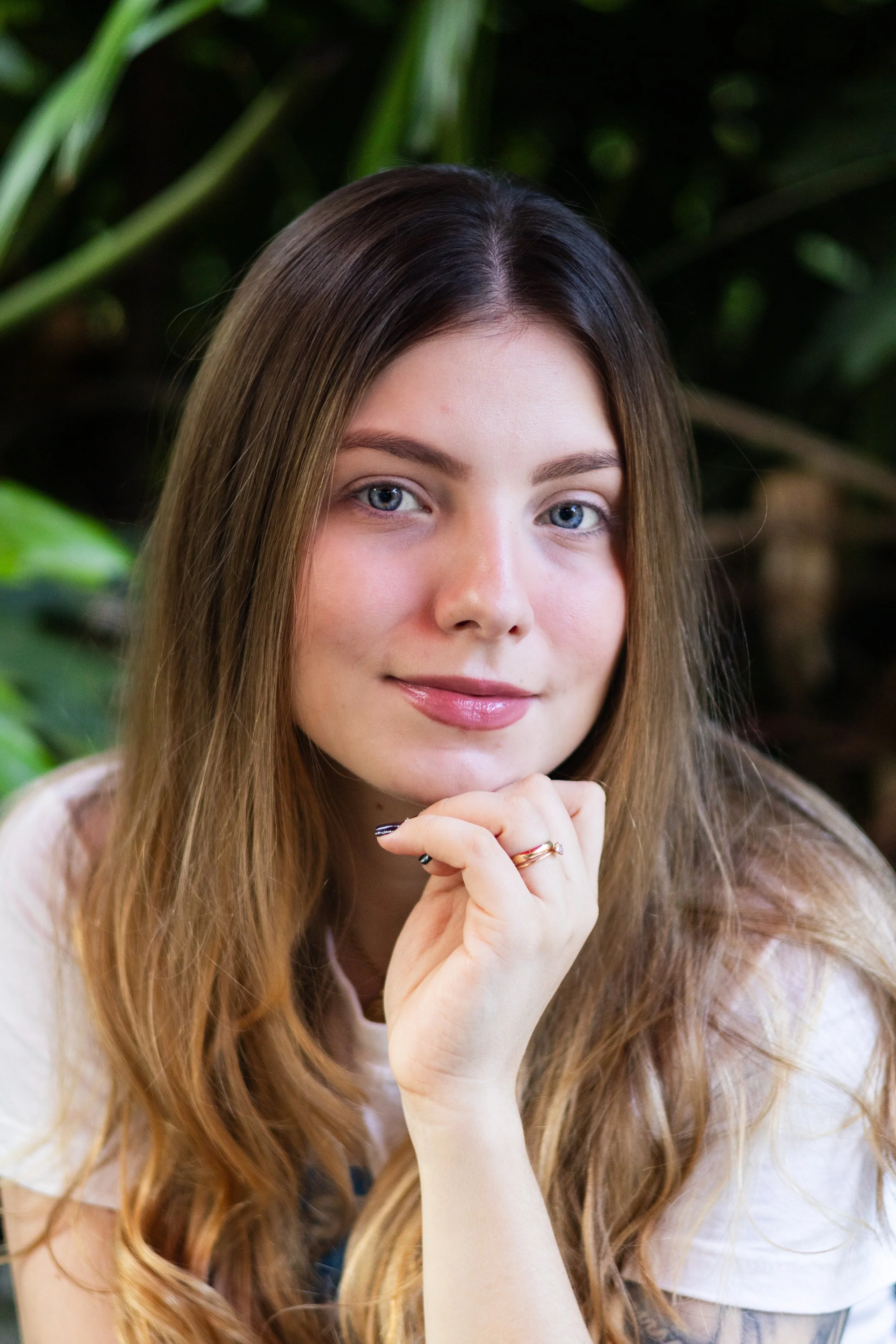Retrato de uma jovem mulher com cabelos longos castanho claro, olhos azuis e pele clara, sorrindo suavemente com a mão apoiada sob o queixo, vestindo uma blusa branca, com fundo de plantas verdes.