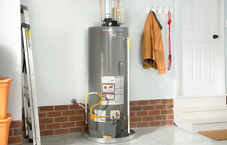 A water heater in a garage with a ladder, jackets hanging on hooks, and steps leading up to a door.